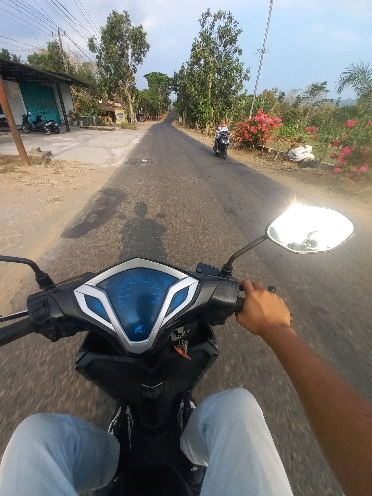 The image is a first-person perspective of someone riding a motorcycle on a road in Ngarjosari, Indonesia. The road is paved and appears to be a rural or semi-rural area. On the left side of the road, there is a building with a corrugated metal roof and a large teal-colored sliding door, with two motorcycles parked in front. Further down the road, there are trees and some houses. On the right side of the road, there are pink flowering bushes, trees, and some white sacks. Another motorcycle is visible further up the road, carrying a person wearing a headscarf. The sky is blue and clear, with some wires and a utility pole visible. The rider's hands are on the handlebars, and their legs are visible in the lower part of the frame, wearing light-colored pants and dark shoes with white stripes. The motorcycle's dashboard with a speedometer is also in view. The sun is shining, casting a shadow of the rider on the road.