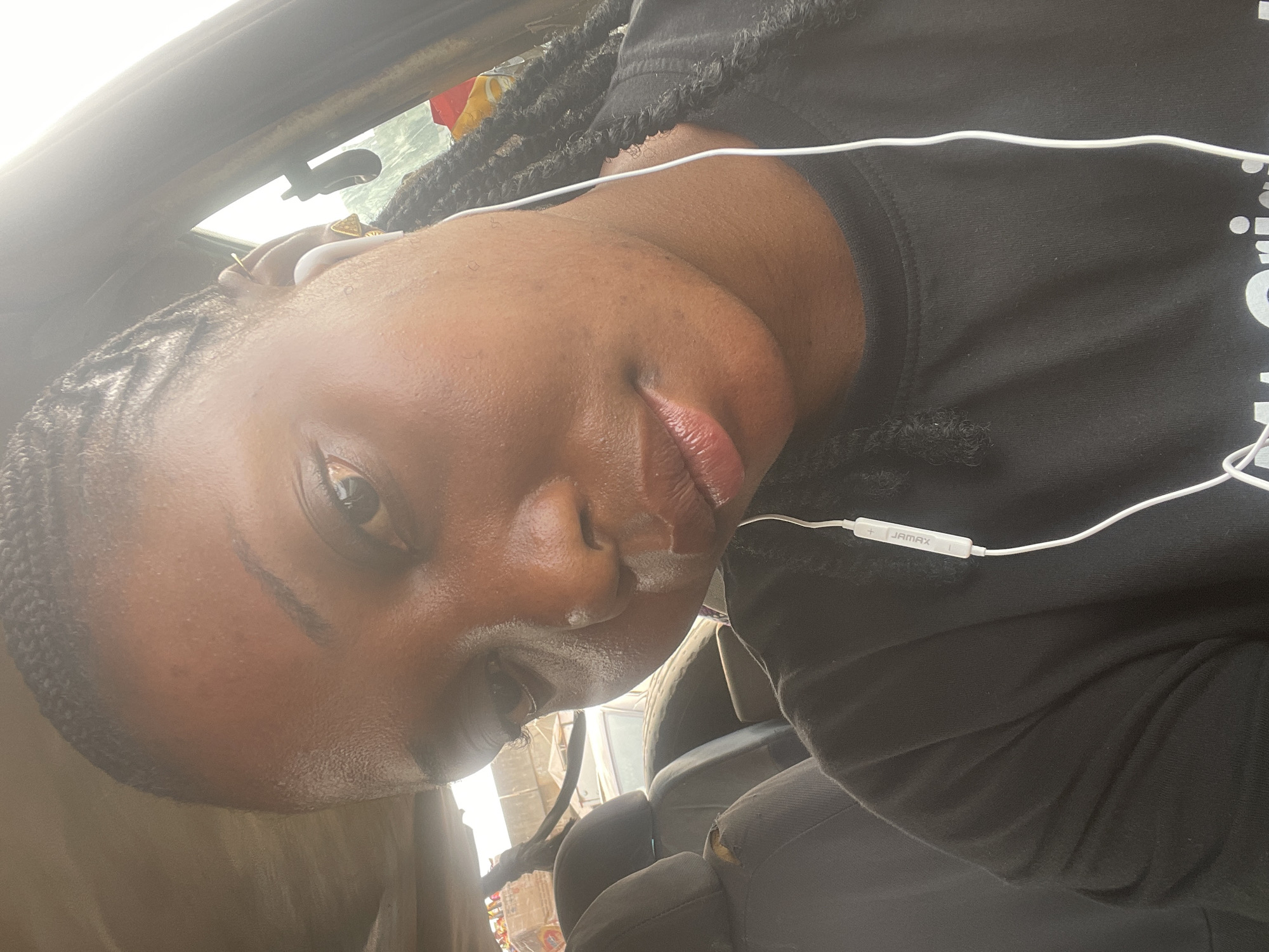 A close-up photograph captures a person with dark skin and braided hair, wearing a black shirt and white earphones. The subject is indoors, likely inside a vehicle, indicated by the visible car interior and reflections on the window. The ambient light suggests daytime. The background details are indistinct, hinting at an urban environment, but not allowing the determination of the city or location of Nyanya, Nigeria.
