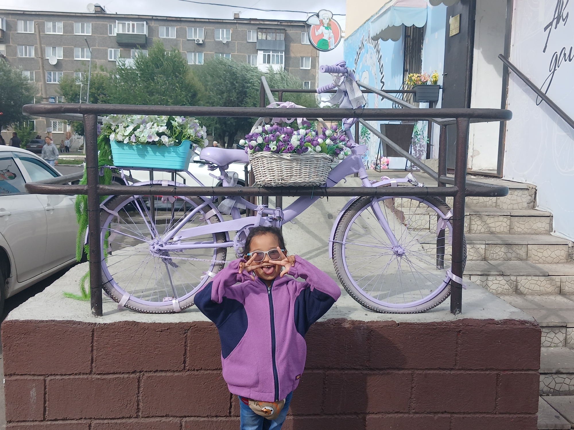 In Щучинск қаласы әкімдігі, Kazakhstan, a young girl stands in front of a purple bicycle adorned with flower baskets. She is wearing a purple and navy zip-up jacket, blue jeans, and a tan fanny pack. The girl is making a peace sign with her fingers over her eyes and sticking out her tongue. She is positioned in front of a brick wall, and the bicycle is placed on a concrete platform. Behind the bicycle, a building with multiple windows and balconies is visible, along with trees. To the right of the building, stairs lead up to an entrance. The scene appears to be on a street or sidewalk in a town.