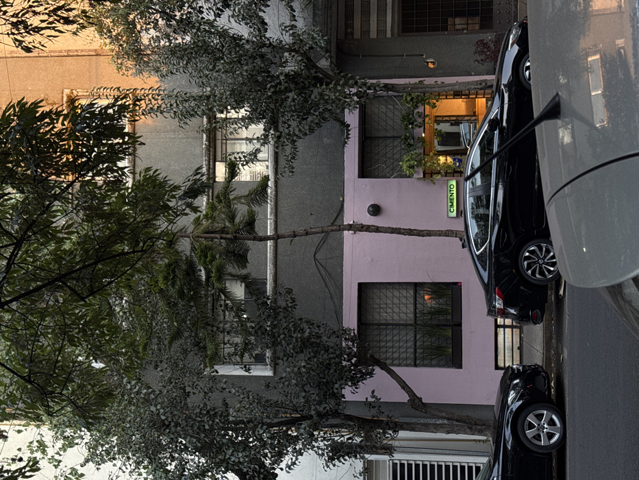 A multi-story building is seen with gray and lavender sections. Windows are visible on each floor. A tree with dense foliage partially obscures the view. A green sign reading "CIMENTO" vertically is attached to the building. Two black cars are parked on the street in front of the building. A portion of a white car is also visible in the right of the frame.