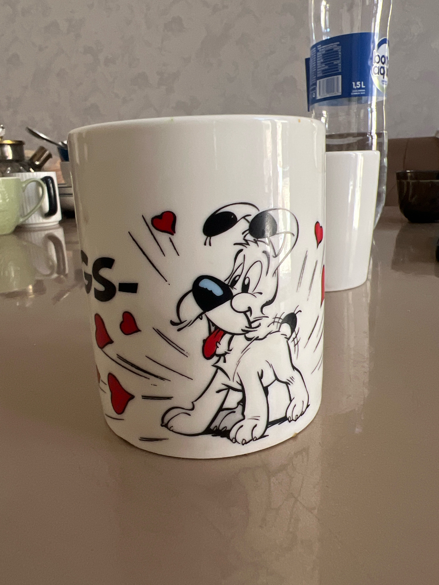 A white mug with a cartoon dog and red hearts is on a light brown reflective surface. The dog is black and white with a red tongue and blue eyes. The text "GS-" is visible on the left side of the mug. In the background, a clear plastic water bottle with a blue label that says "1.5L" is present. To the left of the mug, there are other white mugs and kitchen utensils. The wall in the background has a textured, light gray pattern.