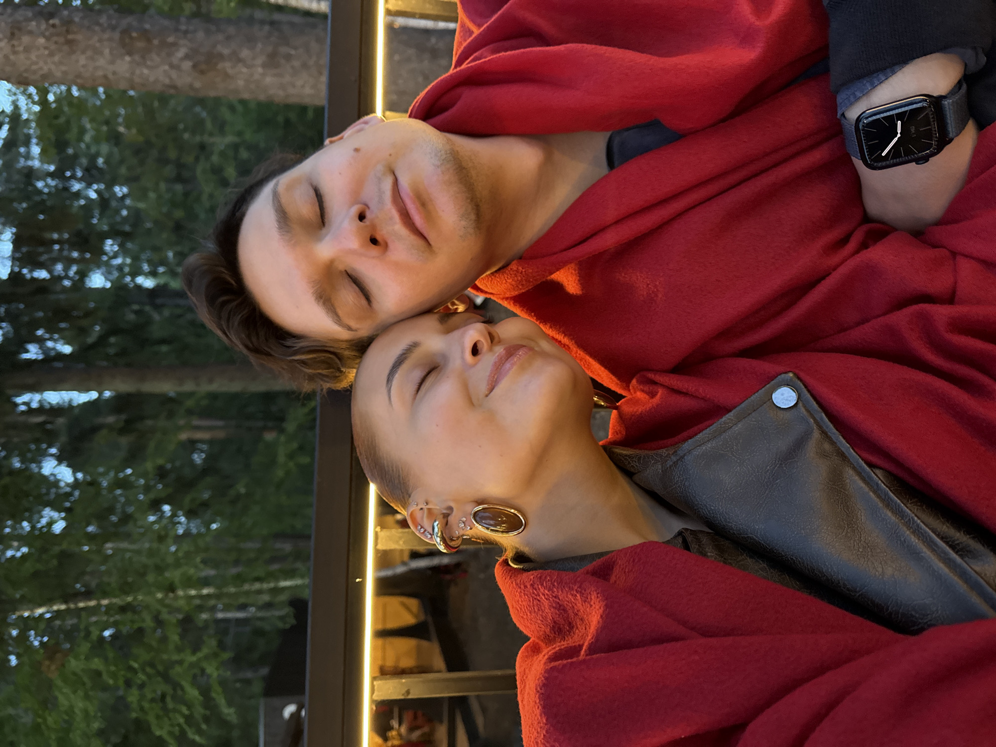 This image, captured in Izhevsk, Russia, depicts an intimate and serene moment between a man and a woman, likely a couple. The original photograph is oriented on its side, but for clarity, the description assumes a portrait orientation. Both individuals have their eyes closed, resting their heads close together, conveying a sense of peace, comfort, and affection.
They are both wrapped in a vibrant red blanket or shawl. The man, on the left, has dark hair and a light stubble. He wears a dark long-sleeved top under the red covering. On his left wrist, he sports a black, square-faced digital watch with a dark strap. The watch face shows white hands against a black background, and small white text "08-41" is visible, likely indicating the time.
The woman, on the right, has light skin and her hair pulled back. She exhibits multiple ear piercings, including a large, oval, dark brown or amber-colored earlobe plug and several smaller, metallic (silver-colored) hoop and stud piercings along her upper ear cartilage. Beneath the red wrap, she is wearing a dark garment that appears to be made of a textured material, possibly faux-leather, featuring a silver-colored snap button.
The setting appears to be outdoors or a semi-enclosed space, like a patio or balcony, during the evening or night. The background features blurred dark green foliage, suggesting trees, and a prominent vertical strip of warm artificial light illuminates the subjects, casting a gentle glow on their faces. The overall atmosphere is one of quiet intimacy and relaxation.
