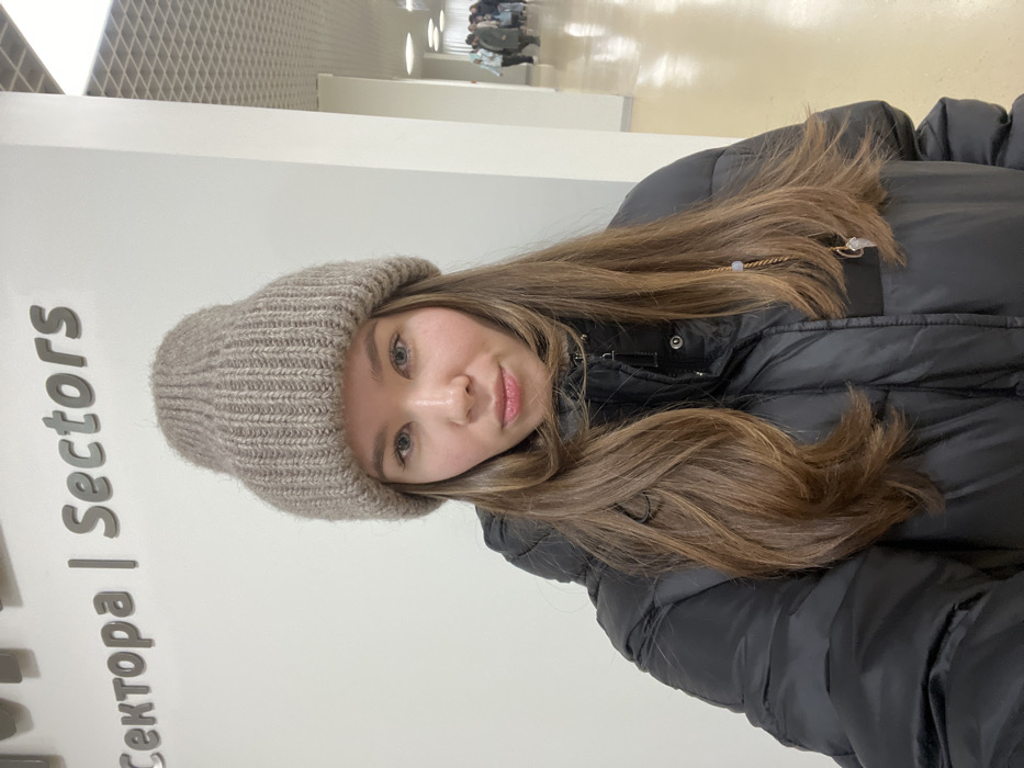 A young woman is featured prominently in a selfie, positioned with her head and upper torso in the frame. Her long, straight to slightly wavy brown hair flows over her shoulders and down her chest. She has fair skin, blue or greenish-blue eyes, and her lips are slightly parted in a faint smile, revealing a natural, almost neutral expression. She is wearing a ribbed, light brown or beige knit beanie hat that covers her ears and forehead. Her attire also includes a dark, likely black, puffer jacket with visible silver-toned snaps or buttons near the collar.
The image appears to be taken indoors, in what looks like a modern public building or facility. Behind the woman is a plain, light-colored wall, which has large, gray, sans-serif text printed on it. The text reads: "сектора | Sectors". The first word, "сектора," is in Cyrillic script, followed by a vertical line separator, and then the English word "Sectors."
In the background, further down a corridor, a few indistinct figures are visible, suggesting other people are present in the area. The floor of the corridor appears somewhat reflective, and the ceiling features a patterned texture, possibly a grid or suspended panels. The lighting is bright and even, consistent with indoor artificial illumination. The image is taken at a slight upward angle and is rotated 90 degrees clockwise.