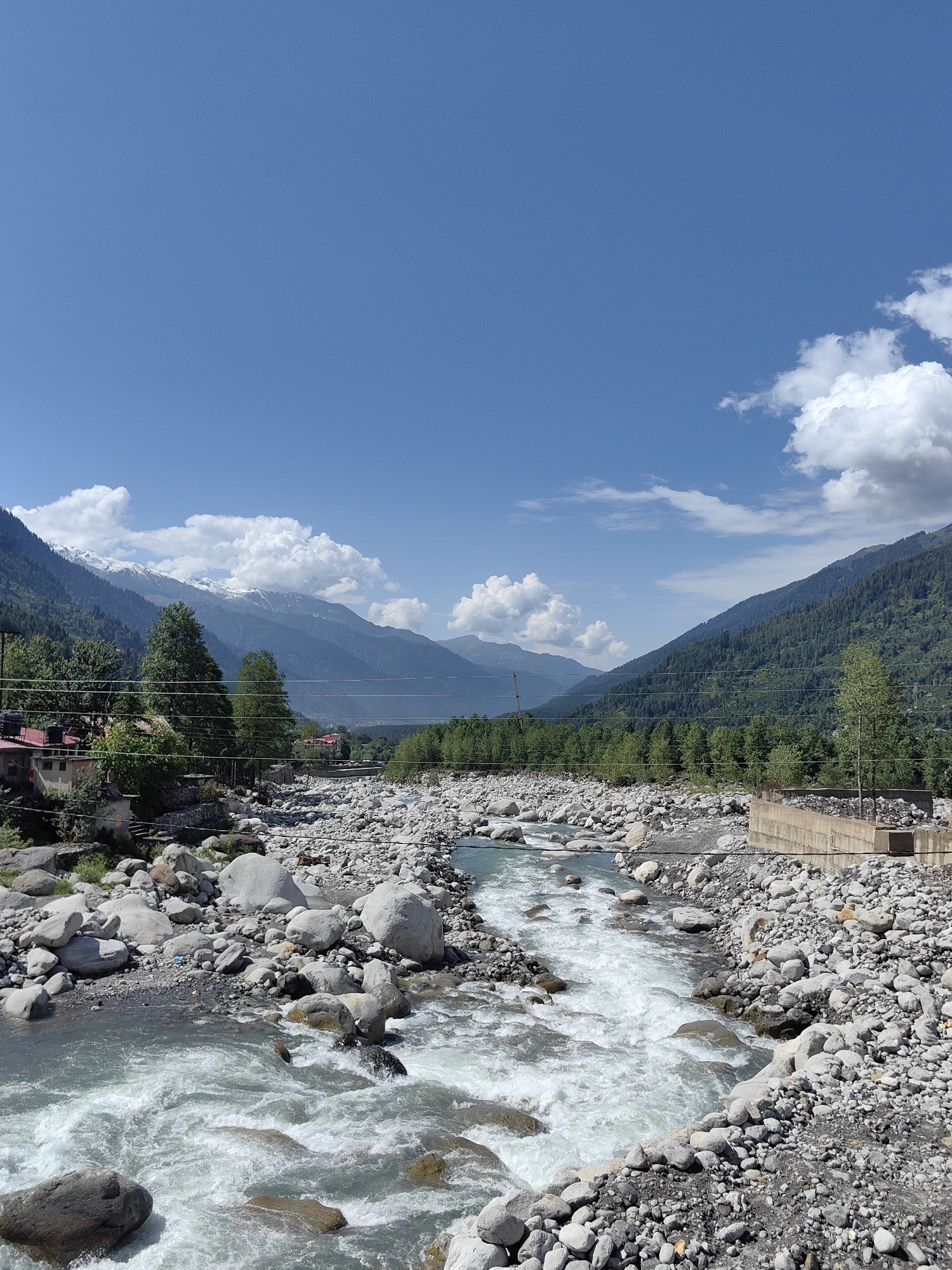The image depicts a wide, shallow river flowing through a rocky terrain, with lush green mountains in the background under a clear blue sky with scattered white clouds. The riverbed is filled with numerous smooth, gray stones of various sizes, and the water is turbulent, with white rapids and foam indicating a moderate current. On the left bank, a few small buildings with red roofs are visible amongst trees, and power lines traverse the scene. On the right bank, a concrete structure, possibly a bridge support or embankment, is partially visible, with more rocks and vegetation. The mountains in the distance are covered in dense green forests, and snow-capped peaks are discernible on the highest ridges. The scene appears to be a natural landscape with some human infrastructure. The location is identified as Vashisht, India.