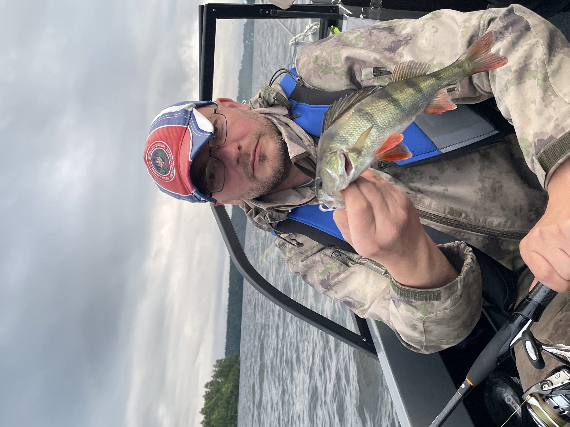 A man in a boat on a body of water is holding up a freshly caught fish. He is wearing a camouflage jacket, a blue and red baseball cap, and glasses. The water is choppy, and the sky is overcast. Trees and a distant shoreline are visible in the background. The boat appears to be a small fishing vessel.
