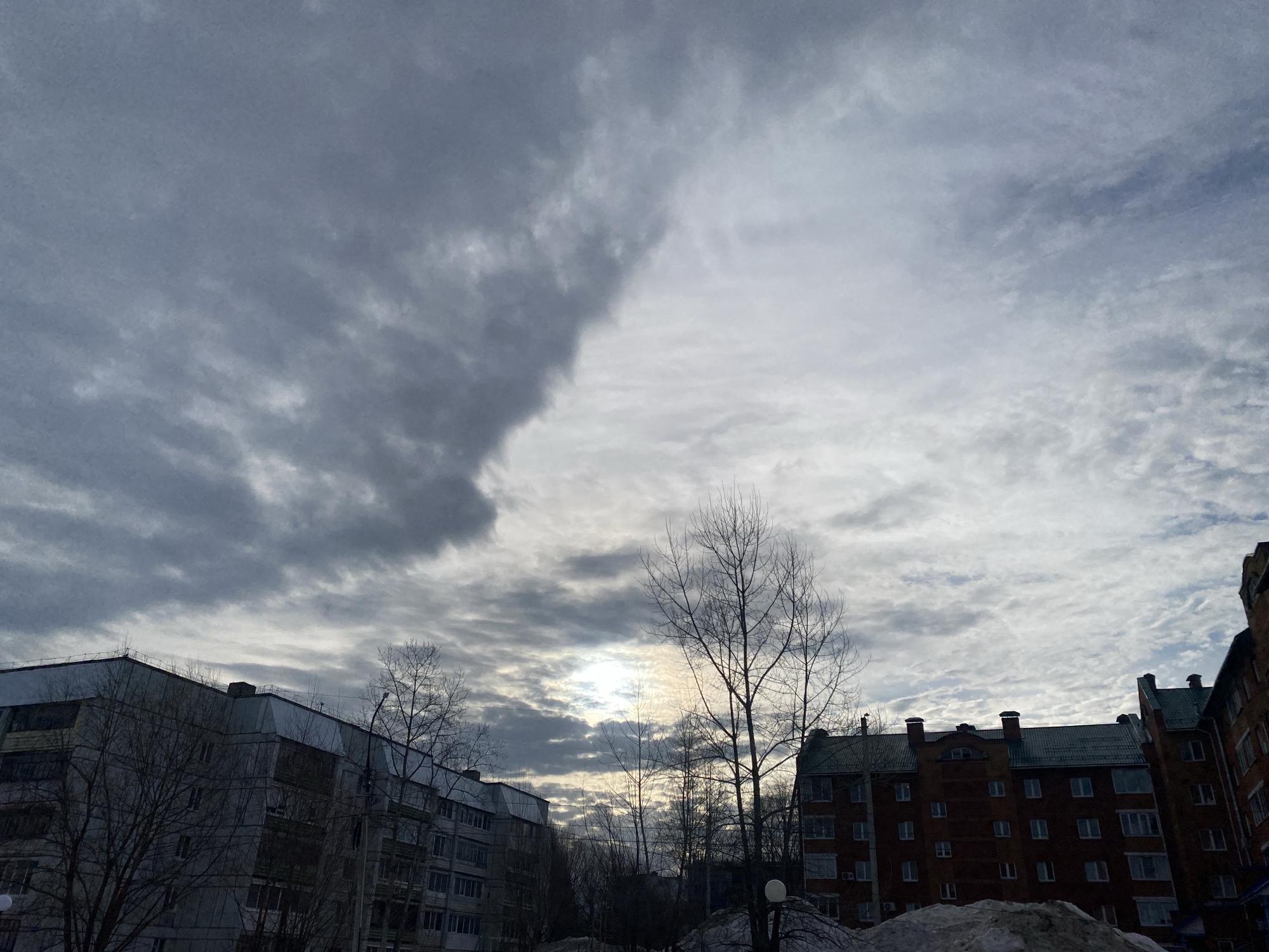 The image presents an urban scene in Chaikovsky, Russia, characterized by overcast skies and the silhouettes of multi-story residential buildings. On the left, a gray concrete apartment building with multiple windows is visible. To the right, a brick building with a dark green roof and decorative elements is situated. Bare trees are scattered throughout the foreground and middle ground, contributing to the wintery atmosphere. Snow piles are visible in front of the buildings. The sky is heavily clouded, with varying shades of gray and a small patch of bright light filtering through. No people are visible in the frame.