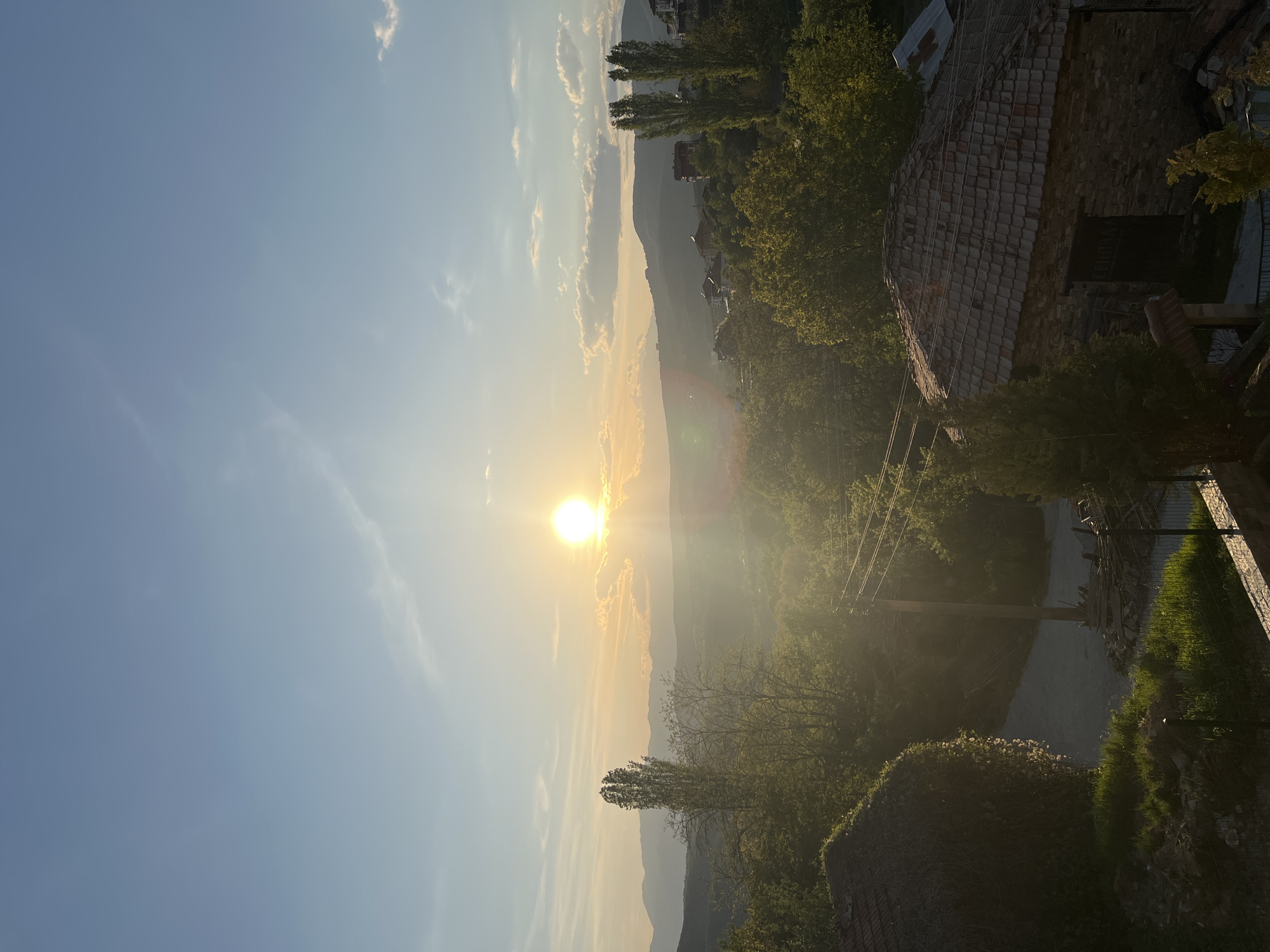 The image shows a landscape in Kribul, Bulgaria, at sunset. The sun is positioned slightly left of center, emitting a strong glare. The sky is light blue transitioning to a pale yellow-orange near the horizon where the sun is located. There are thin, wispy clouds present. In the midground are mountains appearing hazy. The foreground contains trees and what appears to be the corner of a building with a red tiled roof on the right. There are also utility poles and wires visible.