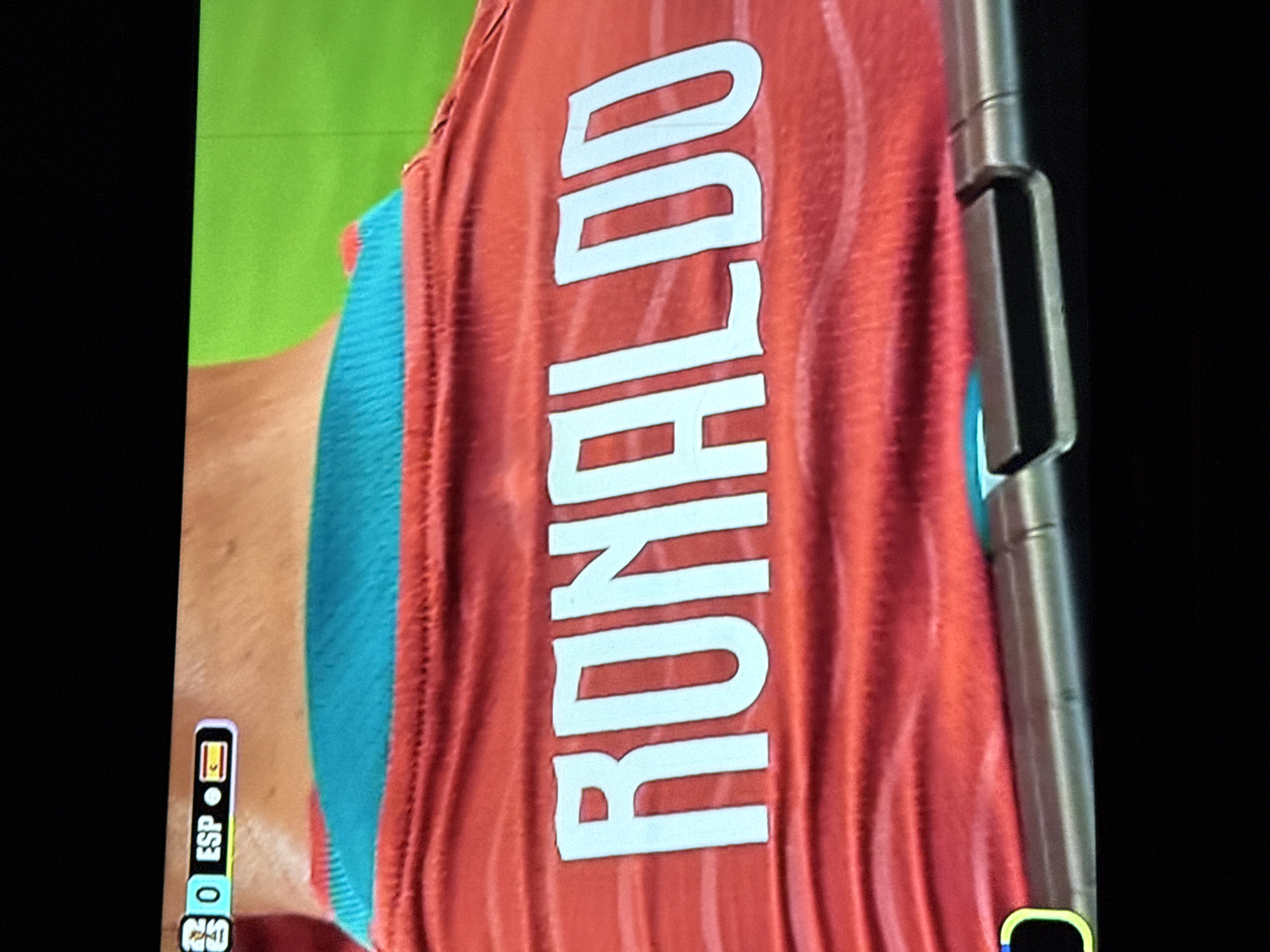 The image shows a close-up view of the back of a red jersey with the name "RONALDO" printed in large white letters. The jersey has a teal colored collar. In the upper left corner of the image, a small graphic displays "0 ESP" followed by the flag of Spain. The jersey is likely a sports uniform, possibly for a soccer match given the name and the scoreboard-like graphic. The image is taken from a screen, as indicated by the slight glare and the quality of the resolution. There is no indication of the city of Ufa, Russia, in the provided image.