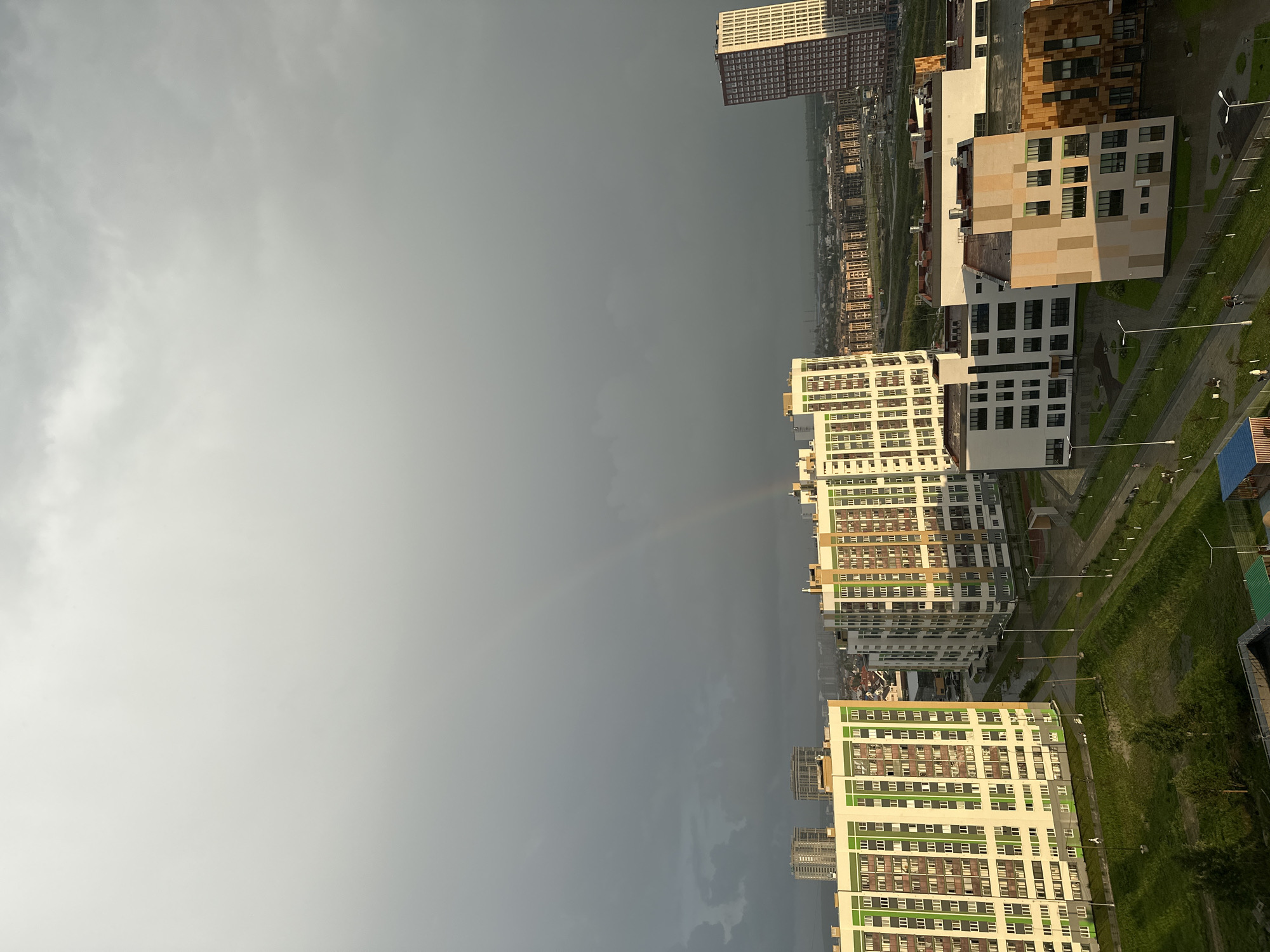 This image depicts an urban landscape in Yekaterinburg, Russia, under a transitioning sky. The upper two-thirds of the frame are dominated by dark, heavy grey storm clouds, with a lighter grey area on the right where a faint rainbow is distinctly visible, arching downwards. Sunlight, likely from the left or rear of the viewer, illuminates the buildings in the lower portion of the image, creating a strong contrast with the dark sky.
In the midground, several multi-story residential buildings are prominent. On the left, a large building features a white facade accented with significant green and some darker grey panels. Adjacent to it in the center, another similar building exhibits white, light brown, and darker grey paneling. To the right, a cluster of shorter, multi-story buildings display varied modern architecture, including white facades with dark windows, and others with checkerboard patterns of beige, light brown, and darker brown panels. On the far right, a tall, slender residential building with a reddish-brown and white patterned facade stands out. In the distant background, a sprawling cityscape with numerous high-rise structures extends to the horizon.
At ground level, well-maintained green spaces, including lawns and some trees, are visible between the buildings. Paved pedestrian pathways wind through these green areas, marked by streetlights. Several individuals are observed walking along these pathways, particularly in the lower-left and lower-right sections of the image. A small structure with a blue roof is partially visible in the bottom-left corner. The overall scene captures the immediate aftermath or a break during a weather event, characterized by the dramatic sky, direct sunlight, and the presence of a rainbow over a modern residential development.