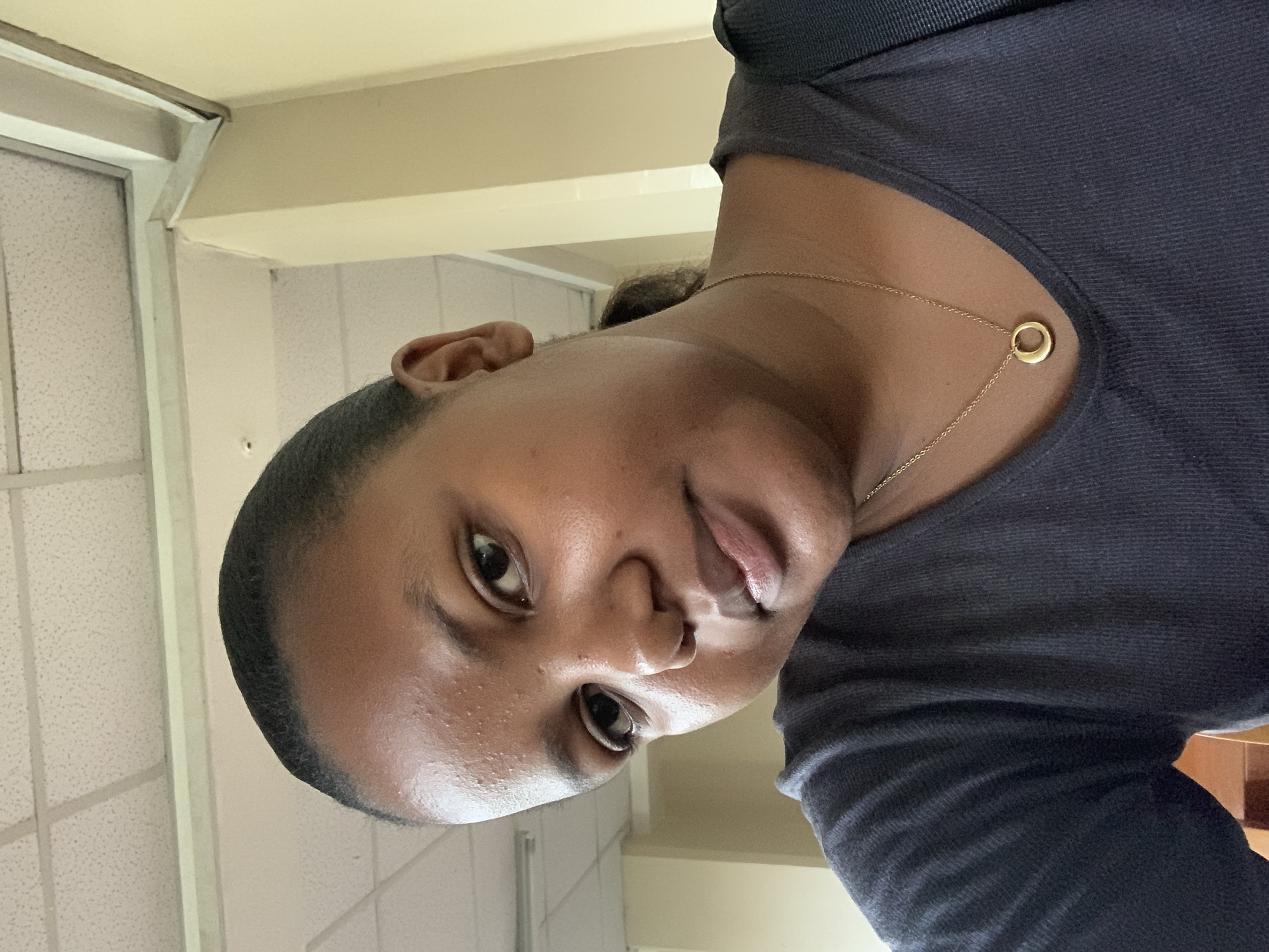 This image is a close-up, tilted selfie of a person, likely a woman, with dark skin, taken indoors in Dar es-Salaam, Tanzania. Her dark hair is pulled back, and she is wearing a dark gray or navy crew-neck top. A delicate gold-colored chain necklace with a small, solid circular pendant is visible around her neck.
The subject is looking directly into the camera with wide, open eyes. Her mouth is slightly agape, revealing a hint of her tongue, which gives her expression a somewhat playful or curious quality. The background indicates an indoor setting, featuring light-colored walls, a window or door frame on the left, and a drop ceiling with acoustic tiles. The lighting appears to be artificial or indoor. No specific time of day, weather conditions, or visible text can be determined from the photograph.