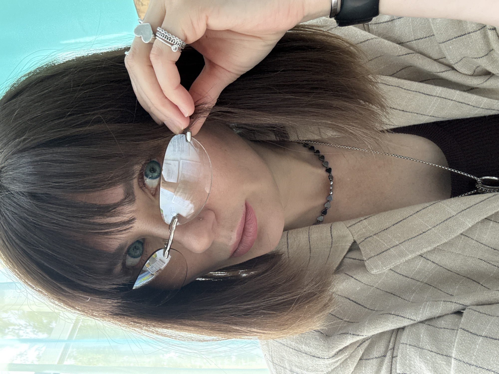 The image shows a close-up, chest-up portrait of a woman with brown hair and bangs, wearing frameless, rimless glasses. She has green eyes and is wearing a pinstripe jacket over a dark top. She is also wearing a beaded necklace and several rings on her right hand, including a heart-shaped ring. The background is blurred, but appears to be an indoor setting with a blue-tinted window. There is no discernible information in the image that would suggest a location in Chaikovsky, Russia.