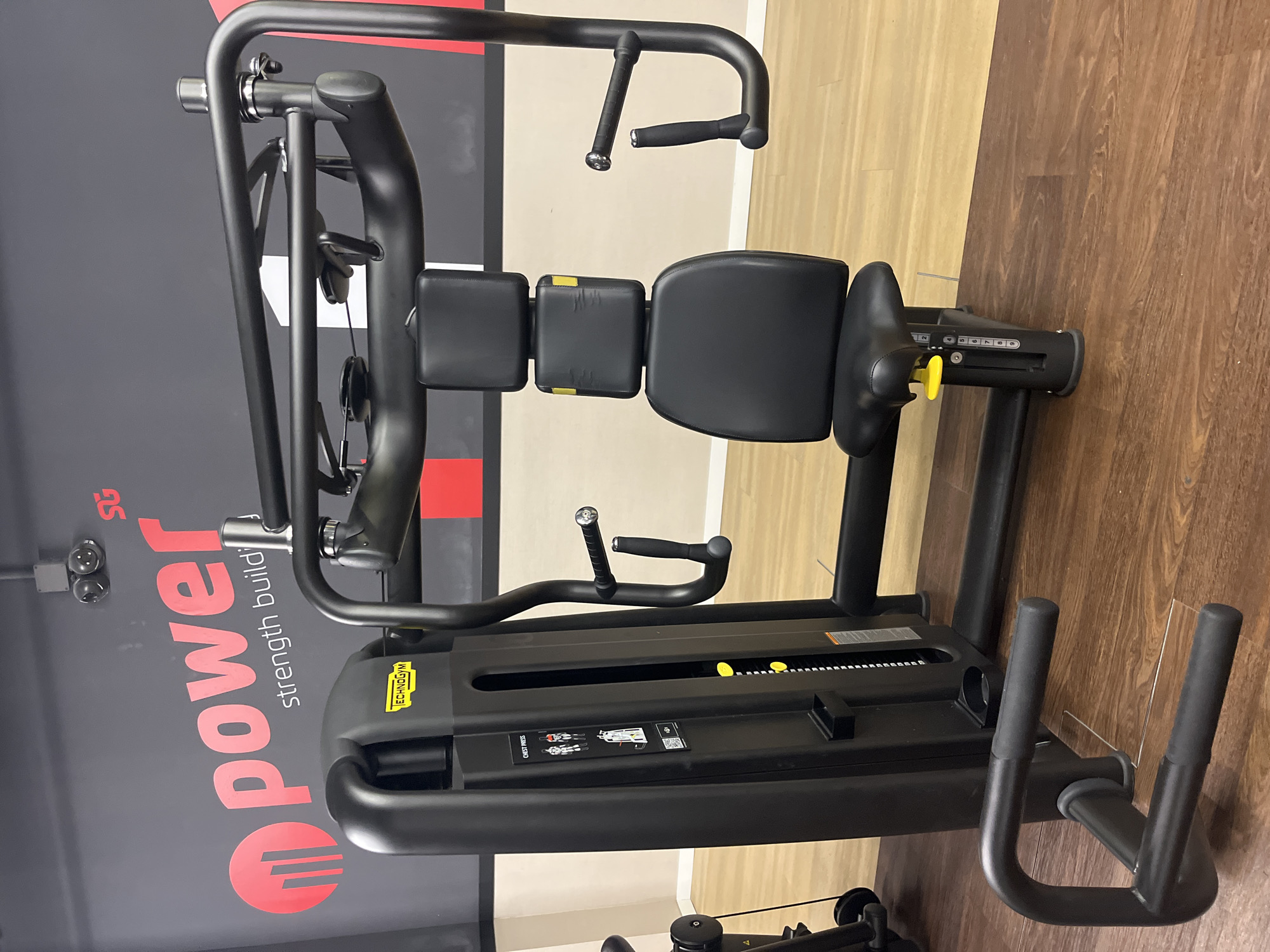 The image shows a chest press machine in a gym setting. The machine is black with yellow accents. It has a padded seat and chest rest. The backdrop includes a sign that says "power strength building." The gym has two types of flooring: light wood and dark wood. The location is a fitness center, and while it's impossible to confirm it is in Barcelona, Spain, based on the image alone, it's a plausible setting for this type of gym equipment. There are no people visible.