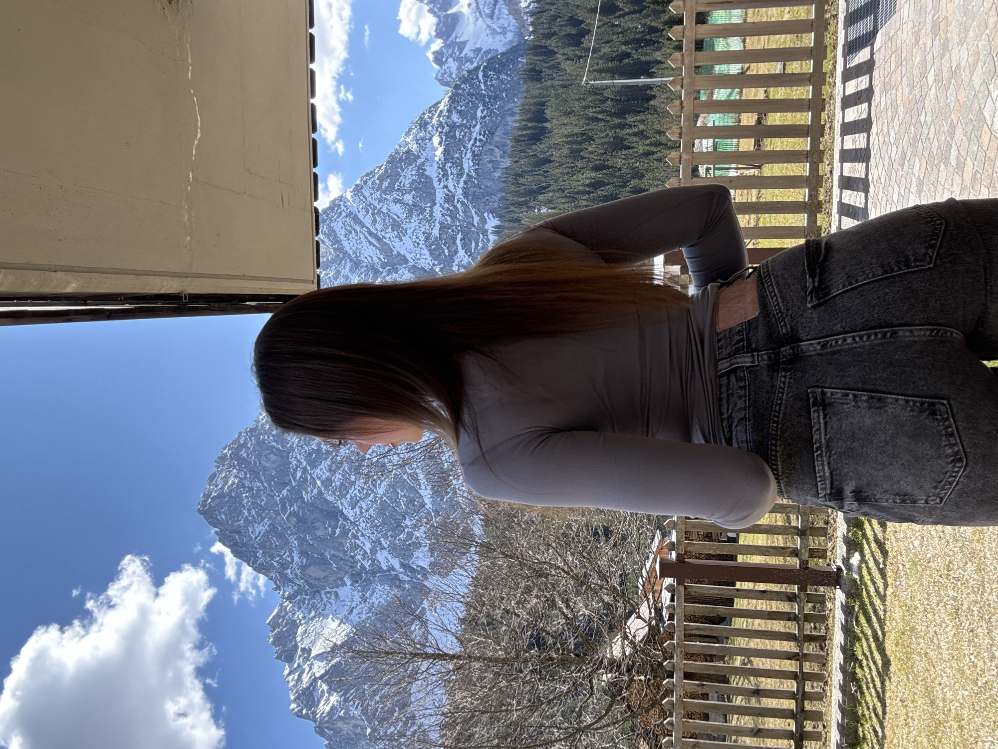 A woman stands on a balcony overlooking a mountain range in Sappada / Plodn / Sapade, Italy. She is wearing a gray long-sleeved shirt and dark jeans. The balcony has a wooden railing and a stone floor. In the background, a snow-covered mountain rises above a dense forest. The sky is blue with scattered clouds. A portion of a building overhangs above the woman.