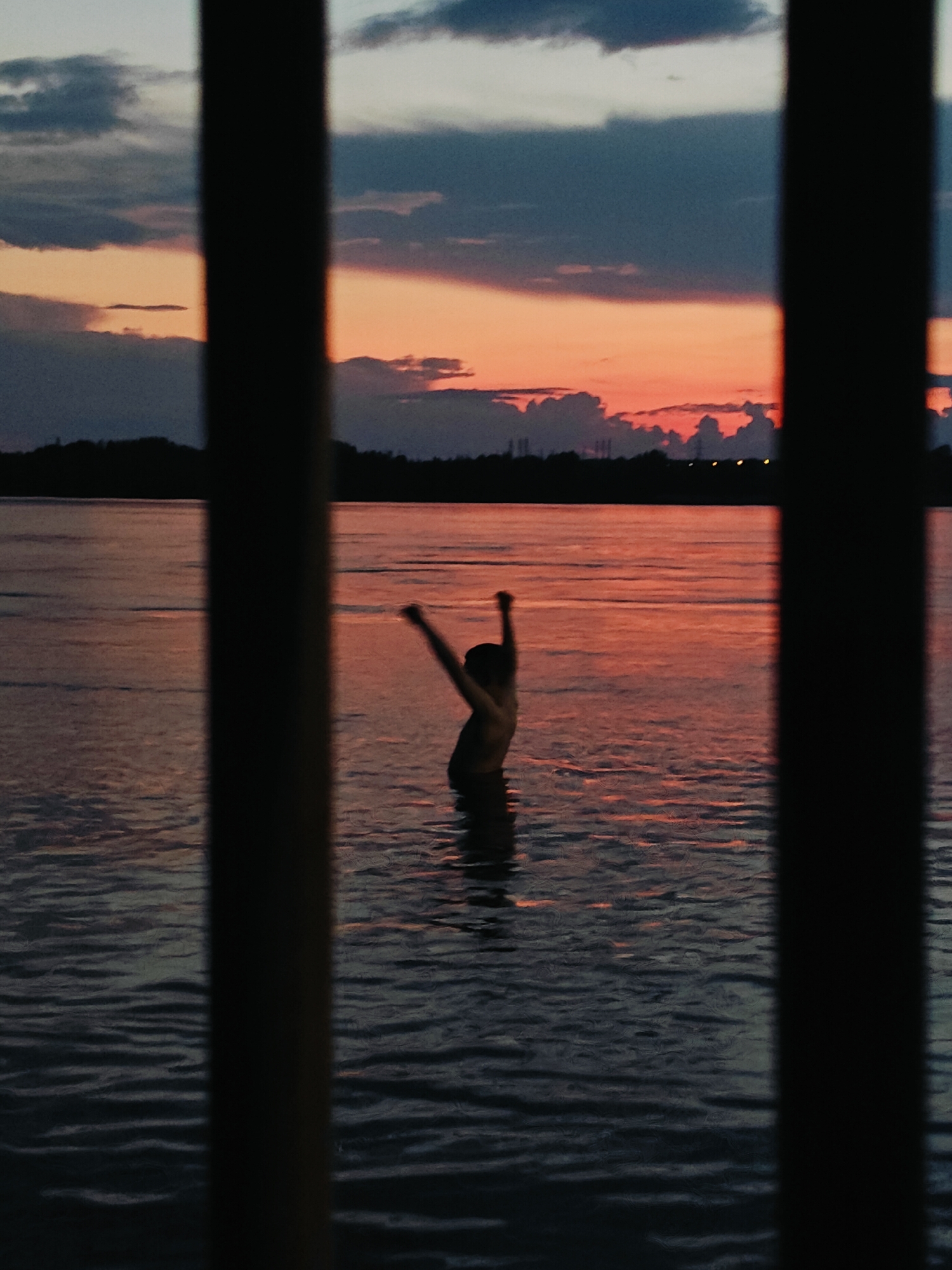 A silhouette of a person stands in the water, with arms raised, against a sunset sky. The sky is a blend of orange, pink, and deep blue clouds. The person is facing away from the camera, and their body is partially submerged in the water. The foreground is dominated by the dark, vertical bars of a fence or railing, which obscure parts of the scene. The water appears calm with ripples reflecting the colors of the sunset. In the distance, a dark tree line or cityscape is visible along the horizon. The overall impression is one of peacefulness and freedom, with the sunset creating a dramatic backdrop.