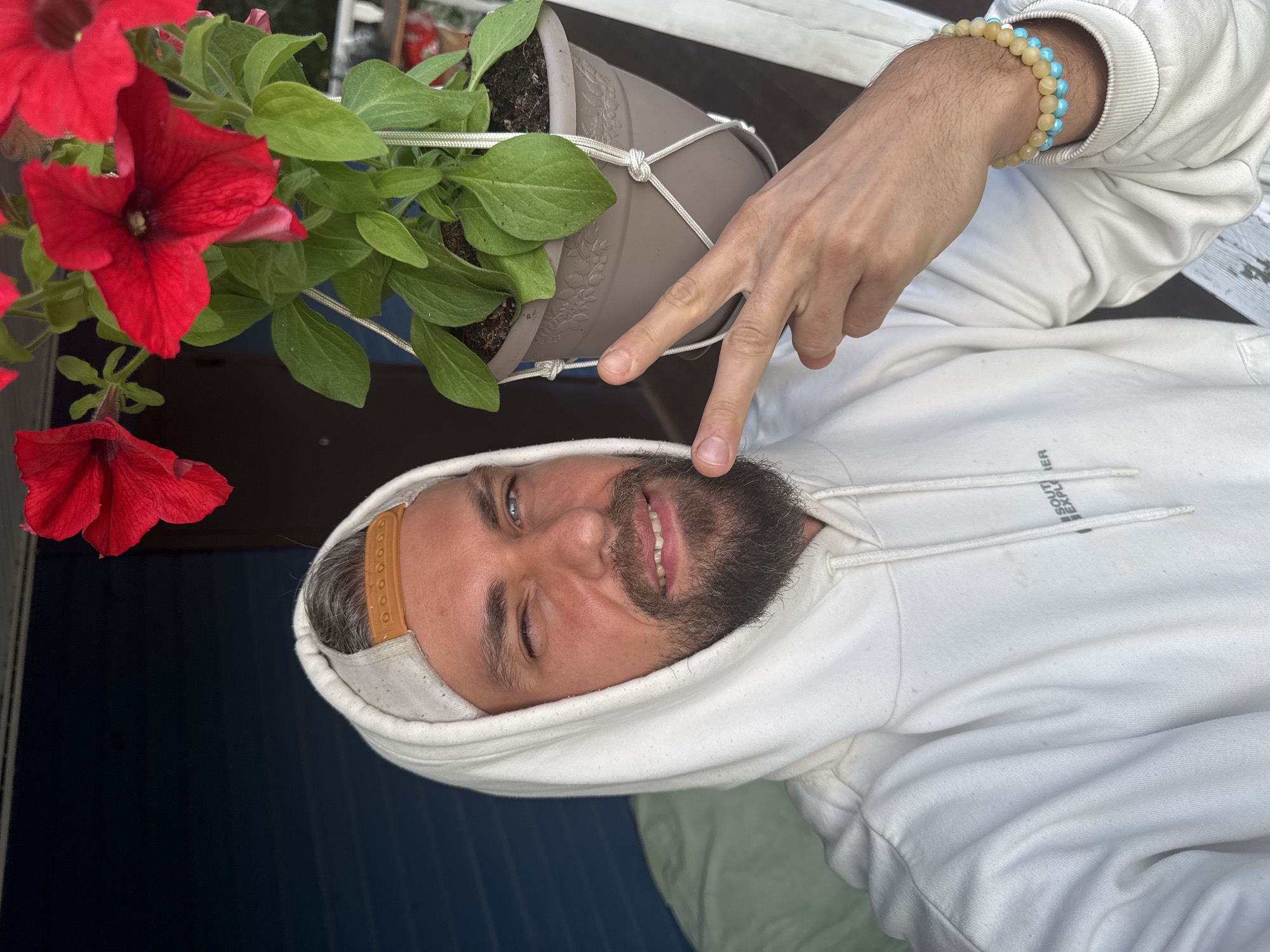 A man with a beard and wearing a white hoodie and a baseball cap is taking a selfie outdoors, with red petunias in a hanging planter to his right. He is winking and holding up two fingers in a peace sign. He is also wearing two bracelets on his left wrist, one with yellow beads and another with blue beads. The background appears to be a blue wooden wall. The image does not provide any information about the location within the city of Kuti, Russia.
