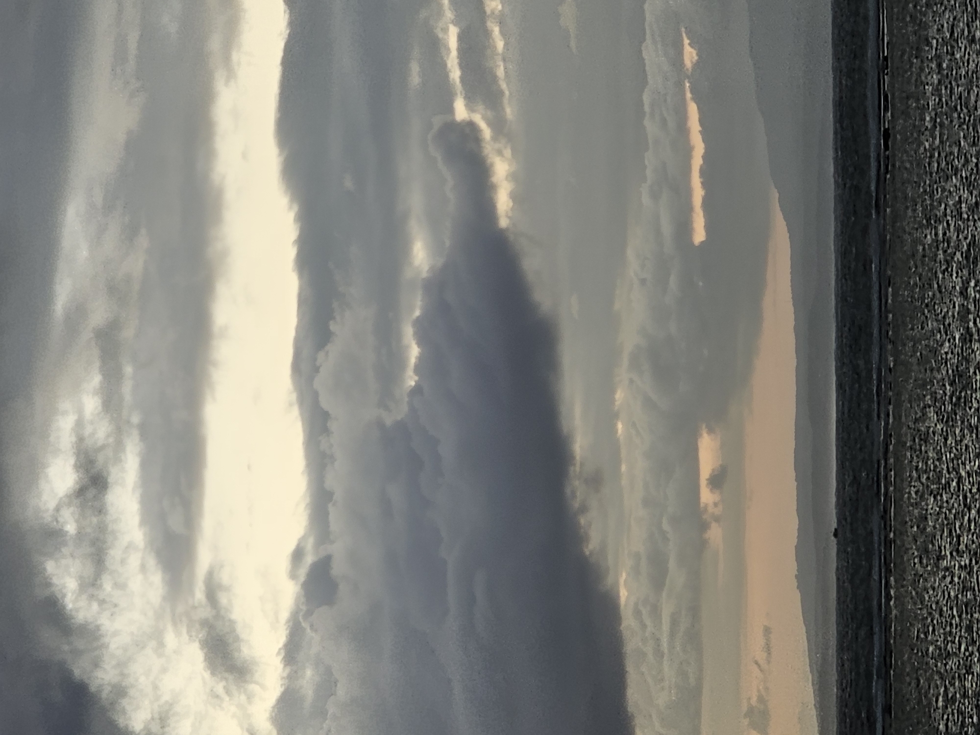 The image depicts a vast, cloudy sky over a body of water, likely the sea. The clouds are a mix of dark grays and lighter, more diffused whites, with patches of pale yellow light suggesting the sun is either setting or rising behind them. The sky occupies the majority of the frame. Below the sky, a hazy line of distant land, appearing as rolling hills or mountains, is visible. In the foreground, a dark, rippling expanse of water stretches across the bottom half of the image. A small, dark boat is barely visible on the water in the middle distance. The overall impression is one of a serene, possibly overcast, coastal scene. The location is not identifiable from the image itself, and there is no information to indicate it is in Denpasar, Indonesia.