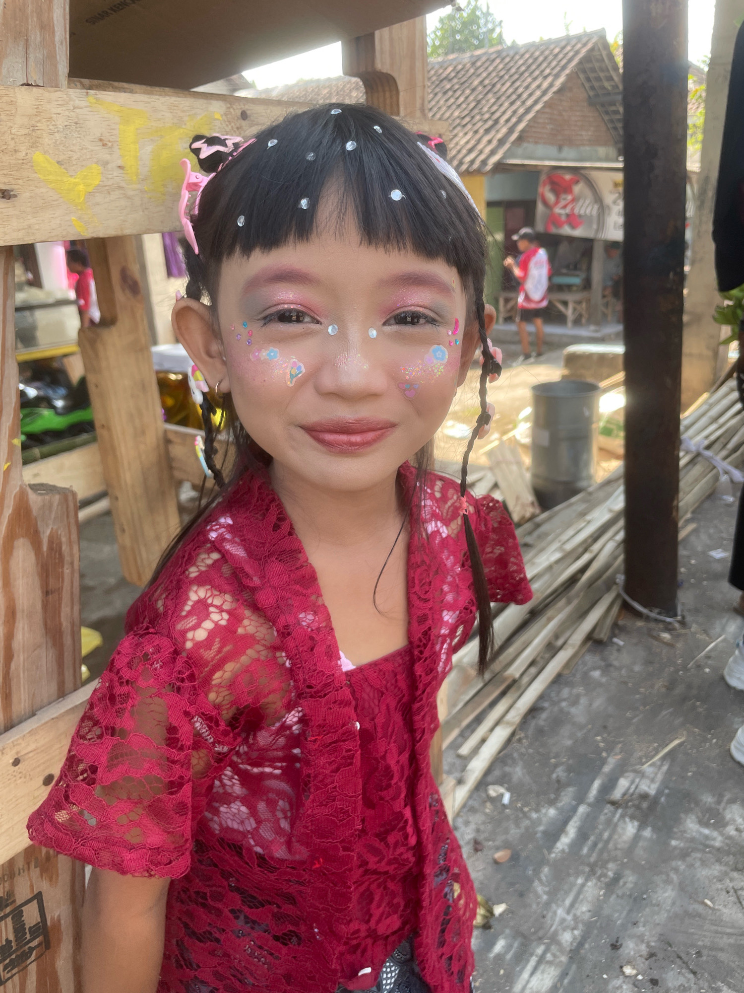An Indonesian girl, adorned with glitter and makeup, stands outdoors. She is wearing a vibrant red lace top and has her hair styled in pigtails. The background suggests a rural or semi-urban setting, possibly in Kebon Waris, Indonesia, with buildings and what appears to be construction materials. The lighting indicates it is daytime.