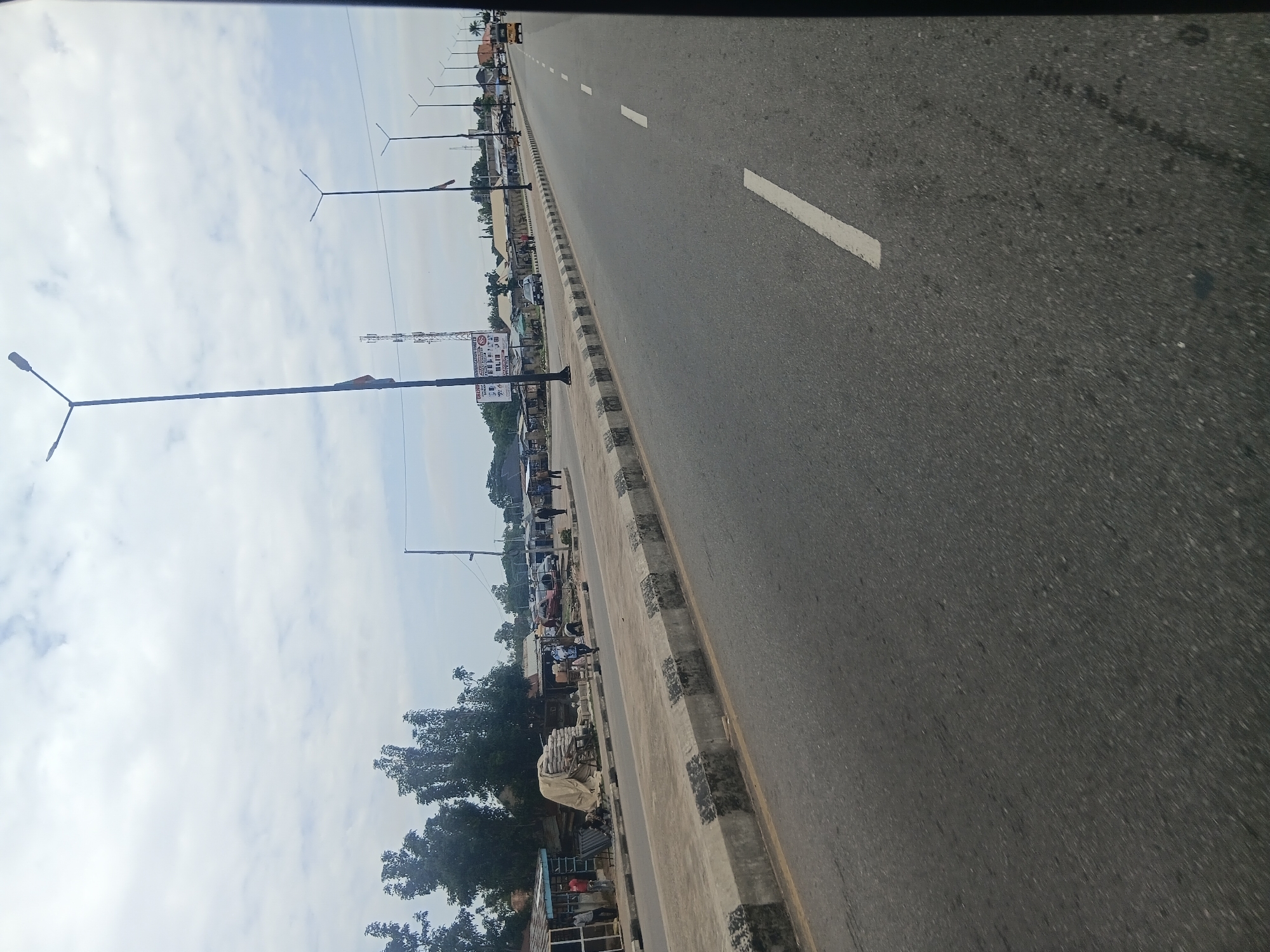 The image shows a street scene in Jalingo, Nigeria. The perspective is from a vehicle moving forward, looking slightly to the left. The road is asphalt with white lane markings. On the left side of the road, there is a concrete median with black and white striped edges. Beyond the median are buildings that appear to be shops and stalls, with some people standing around. Trees are visible behind the buildings. Several streetlights are present along the roadside. In the distance, there are more buildings and trees, and a cell tower. A yellow trishaw is visible on the road in the far distance. The sky is overcast with clouds.