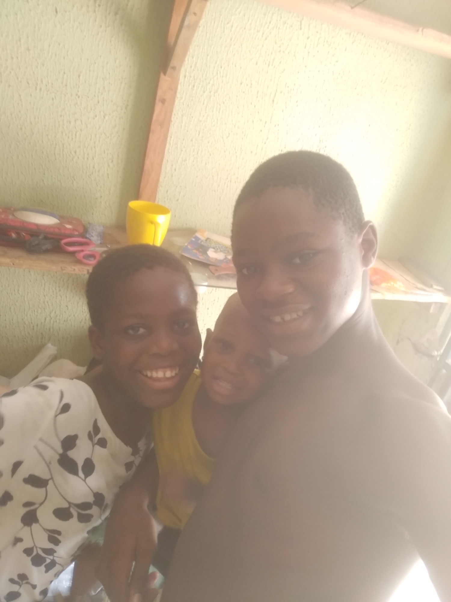 A bright, daytime photograph captures three children, likely boys, positioned closely together and smiling warmly at the camera. The child on the left wears a white top adorned with a black leaf or floral pattern and displays a wide, cheerful smile. Partially visible behind and to their right is a second child, wearing a yellow top and also smiling. The third child, on the far right, is bare-chested and grins broadly. This child's face and upper body are significantly overexposed due to a strong light source originating from the right side of the frame.
The background reveals an indoor setting, likely a home. A light green or yellowish-green wall with a textured finish is visible. On a wooden shelf to the left, various items are arranged, including a red object (possibly a toy or container), pink scissors, and a vibrant yellow cup. Further to the right, another shelf, potentially made of glass, holds some papers or books. A diagonal wooden beam or support structure can also be seen in the background. The children's expressions convey happiness and camaraderie. No text is visible in the image.