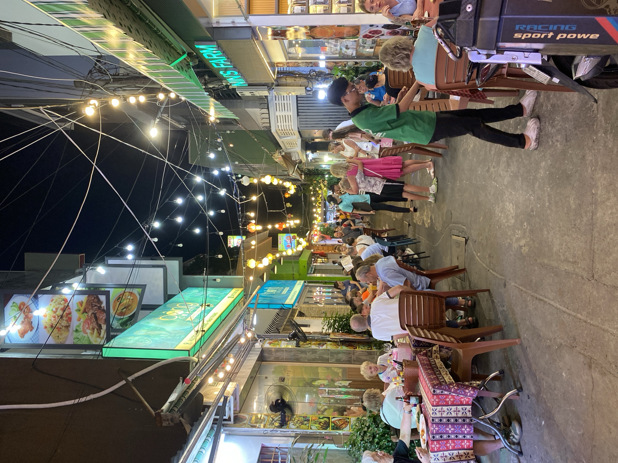 The image depicts a vibrant street scene at night in Nha Trang Ward, Vietnam, likely a pedestrianized area or an alleyway frequented by tourists and locals. Numerous strings of bare incandescent bulbs are strung overhead, illuminating the narrow street and creating a festive atmosphere. The street is lined with restaurants and shops, many of which feature illuminated signage.
On the left side of the image, a restaurant displays a menu with images of food items, including what appears to be fried chicken, rice dishes, and a curry. Diners are seated at tables covered with patterned tablecloths, some engaged in conversation and eating. Various objects are visible on the tables, such as bottles, condiment containers, and plates of food.
On the right side of the street, another establishment, possibly a shop, has signage that reads "MINIS FARM" in illuminated green letters. More diners are seated at tables outside this establishment.
A diverse group of people are present in the scene, including couples, families, and individuals. Some are seated at tables, while others are walking through the street. A man in a green shirt and black pants, possibly a waiter or staff member, is walking through the crowd, holding a tray or bag. Other individuals are seen in various states of activity, such as talking, eating, and walking. The ground is paved with concrete, showing some wear and tear. The overall impression is a lively and bustling evening in a popular dining and shopping area.
