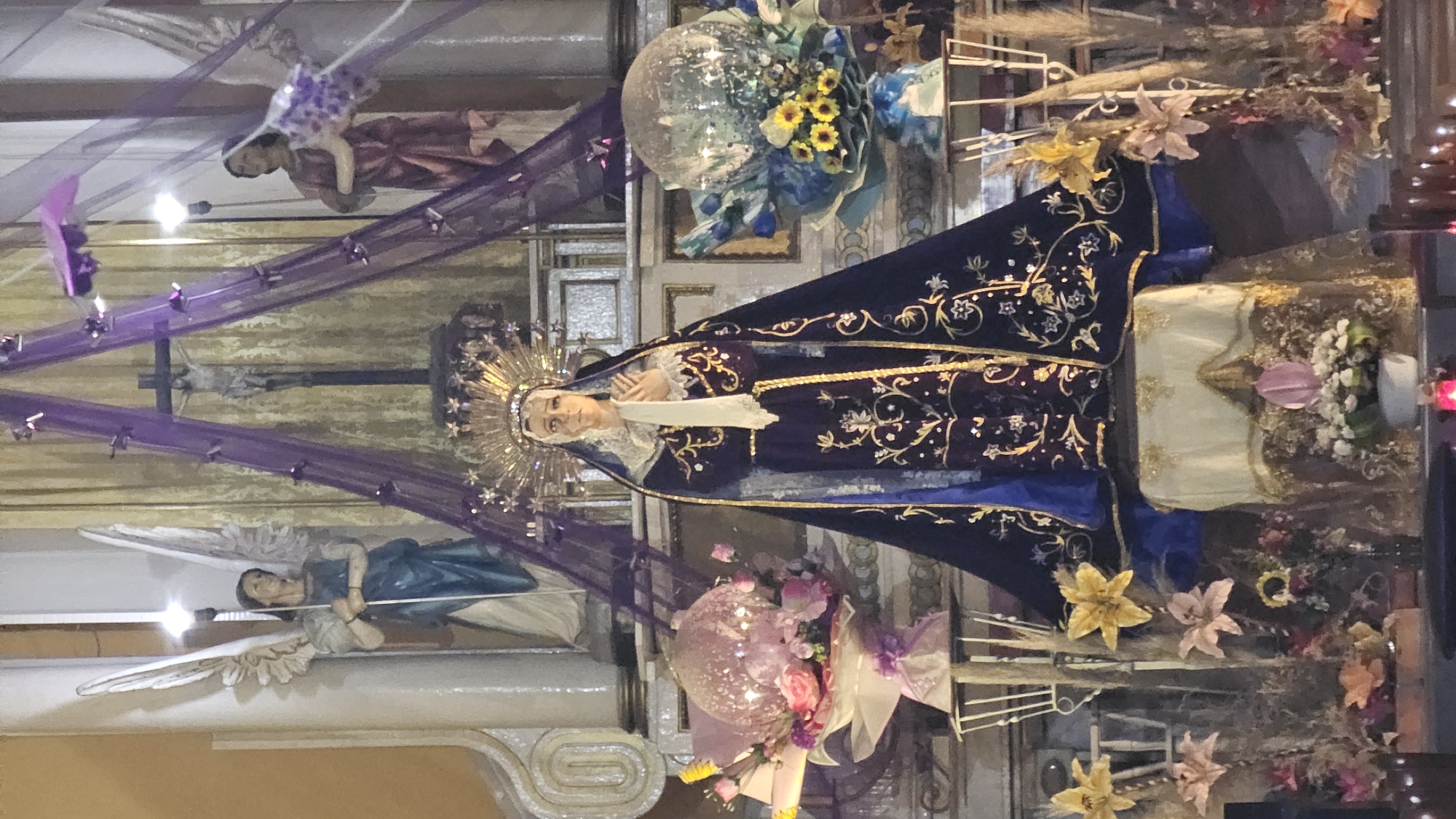 This image captures an elaborate religious display within the interior of a church or chapel, characteristic of Catholic traditions, likely in Puebla, Mexico. The central and most prominent subject is an upright statue of the Virgin Mary. She is depicted wearing a majestic, dark blue robe richly embroidered with gold patterns, over a white undergarment, and has her hands clasped in front of her. Her head is encircled by a radiant gold halo or crown adorned with starbursts.
The scene is highly decorated with various objects and embellishments. Draped throughout the display are swathes of sheer purple fabric, some dotted with star-shaped sequins, adding a festive yet reverent touch. Two statues of angels are visible: one in blue/grey on the lower left, and another in a reddish-brown garment on the upper left, both seemingly suspended. Behind the Virgin Mary, a crucifix with a figure of Christ is partially visible, while to the right, an ornate gold monstrance featuring a radiating sunburst design is prominently displayed.
Abundant floral arrangements enhance the setting. These include large, lush bouquets with blue and yellow flowers, pink roses, and artificial lilies in various hues, often accompanied by glittery or disco ball-like spheres. The Virgin Mary statue stands on or in front of an altar-like structure draped with white and gold fabric. The overall ambiance suggests a special devotional period or a significant religious celebration, such as a Marian feast day. The indoor lighting highlights the intricate details and vibrant colors of the decorations. No discernible text is visible in the image.