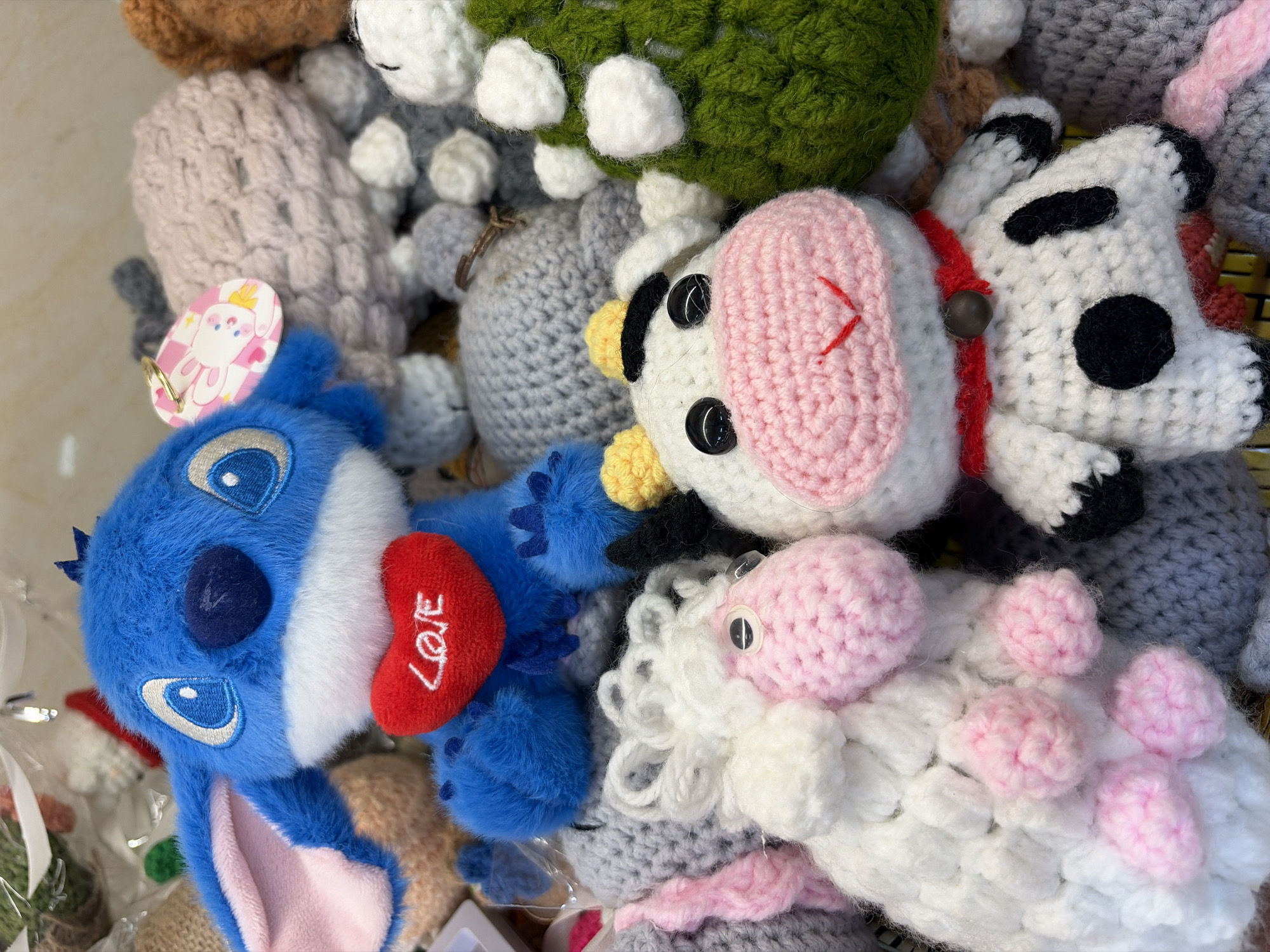 The image shows a collection of crocheted stuffed animals, primarily featuring a blue Stitch character holding a red heart with the word "LOVE" embroidered on it. To the left of Stitch is a white sheep with pink accents, and to its right is a black and white cow with pink accents. Other crocheted items, including what appears to be a green Christmas tree and various grey and pink textured objects, are also visible in the background. The items are densely packed together, suggesting they are displayed for sale or as part of a collection. No specific location in Đà Nẵng, Vietnam, can be identified from the image.