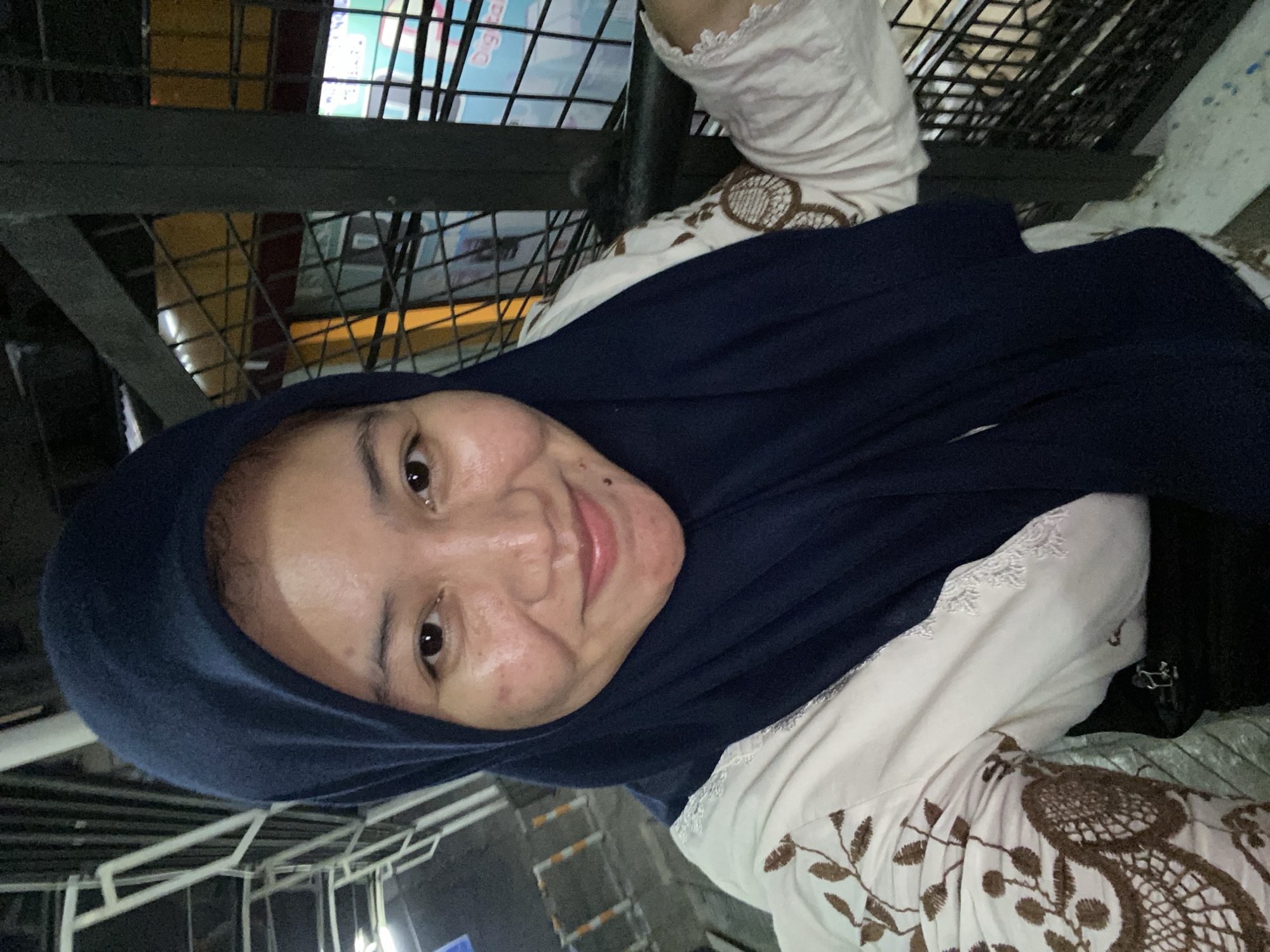 A medium shot photograph features a woman in a dark blue hijab and a light-colored, embroidered shirt. She is smiling and looking directly at the camera. Her face is slightly illuminated, showing some blemishes and freckles. The background is indistinct, but appears to be an outdoor urban environment with metal railings and some signage visible. The image was taken in Banjarbaru, Indonesia.