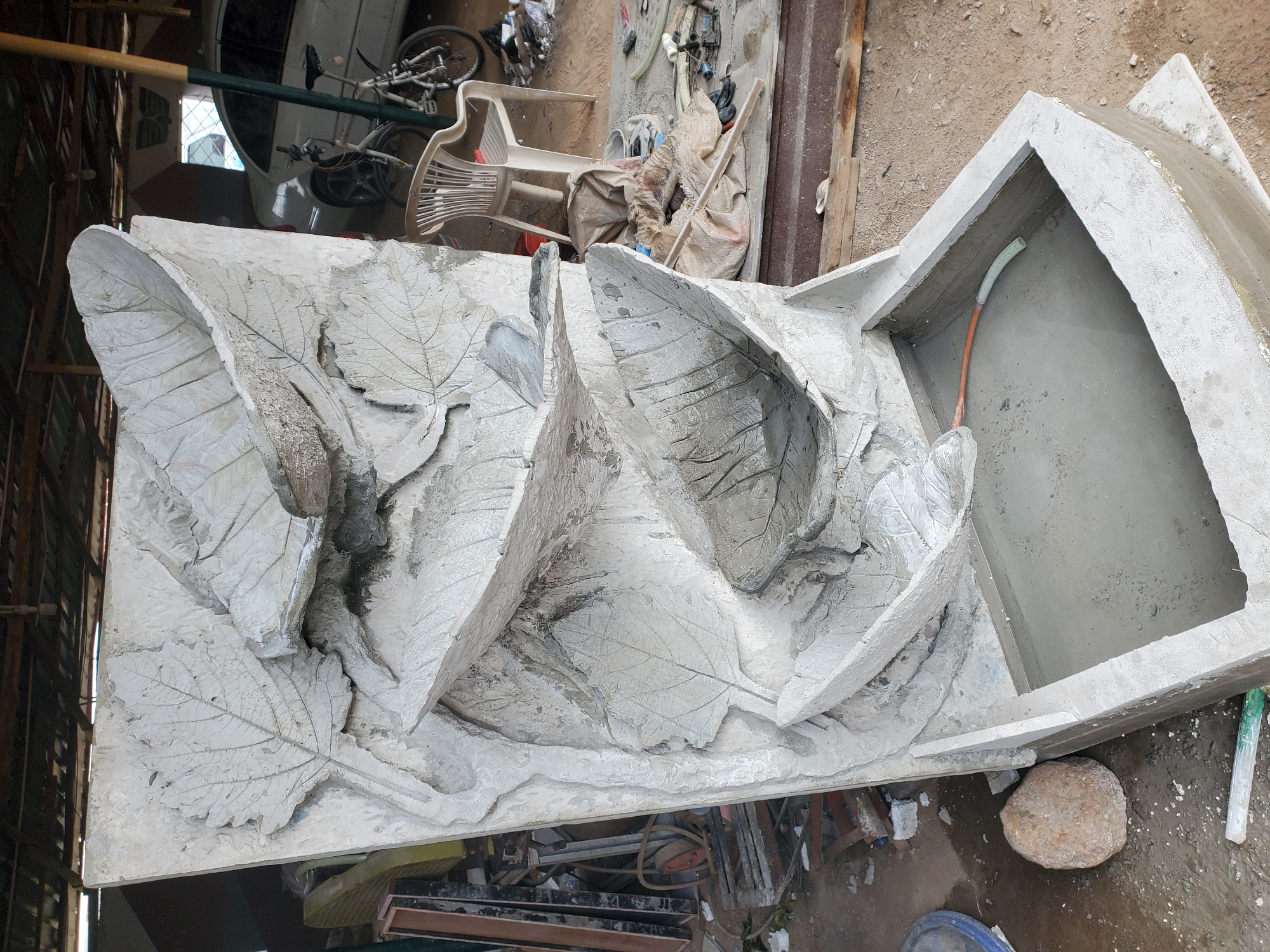 The image depicts a partially constructed concrete water fountain in what appears to be an outdoor workshop or yard in Jos, Nigeria. The fountain is designed to resemble cascading leaves, with three tiers sculpted into the concrete structure, leading down to a rectangular basin. The basin is filled with water and a coiled plastic pipe is visible.
In the background, a white car is parked, and a white bicycle with visible spokes and frame is positioned next to it. A light-colored plastic chair is situated in front of the bicycle. The environment appears cluttered, with various construction materials and tools scattered around, including bags, wood, and what looks like rebar. The ground is a mix of dirt and concrete. The overall impression is of an unfinished project in a somewhat disorganized setting.