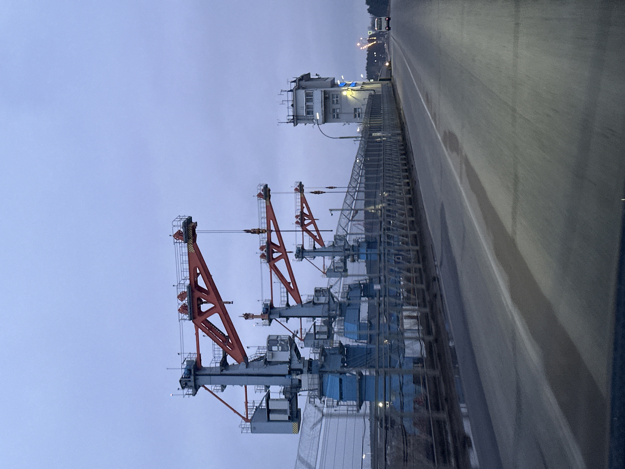 The image depicts a scene along a roadway in Chaikovsky, Russia. A row of large, red and grey container cranes are positioned alongside a fence. Behind the cranes, there is a building with multiple stories, seemingly a control or observation tower. A grey asphalt roadway stretches along the right side of the image. The sky is overcast and grey, suggesting early morning or late evening. There are no visible people.