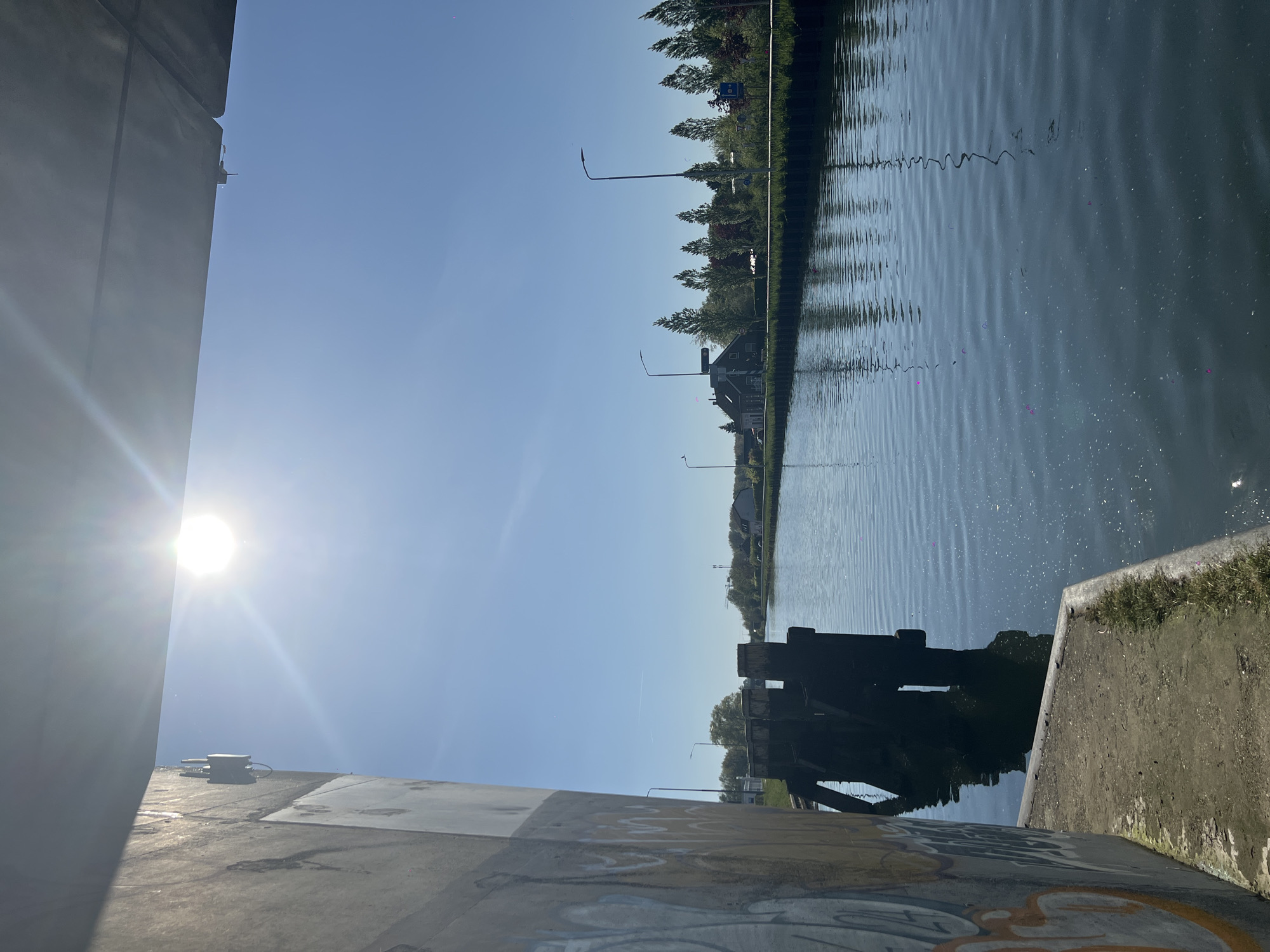The image captures a bright, sunny scene along a canal in Weert, Netherlands, viewed from a slightly elevated position.
In the immediate foreground on the left, a large, imposing concrete structure, likely part of a bridge or embankment, dominates the frame. Its surface is a light grey, showing signs of weathering and some faint, light-colored graffiti patterns near its base. Atop this concrete structure, a small, dark grey box-like object with connected pipes or wires is visible.
The sun, intensely bright, is positioned in the upper left, casting a strong lens flare across the top portion of the foreground concrete and illuminating the clear blue sky.
The main subject is a calm canal or river that stretches horizontally across the mid-ground. Its surface is rippled, reflecting the clear sky and the distant green landscape. On the right side, partially submerged in the water and adjacent to the foreground structure, is a dark, substantial object, possibly a weathered wooden or metal component of a lock gate or a bridge support.
Along the distant right bank, a continuous line of green trees with varied foliage is visible, interspersed with several houses featuring dark roofs and multiple streetlights. A small blue and white sign can be seen among the trees on the far bank.
No specific human or animal activity is depicted; the scene is static and peaceful, bathed in bright daylight, suggesting a clear day in the morning or afternoon. The graffiti on the concrete wall is not clearly legible.