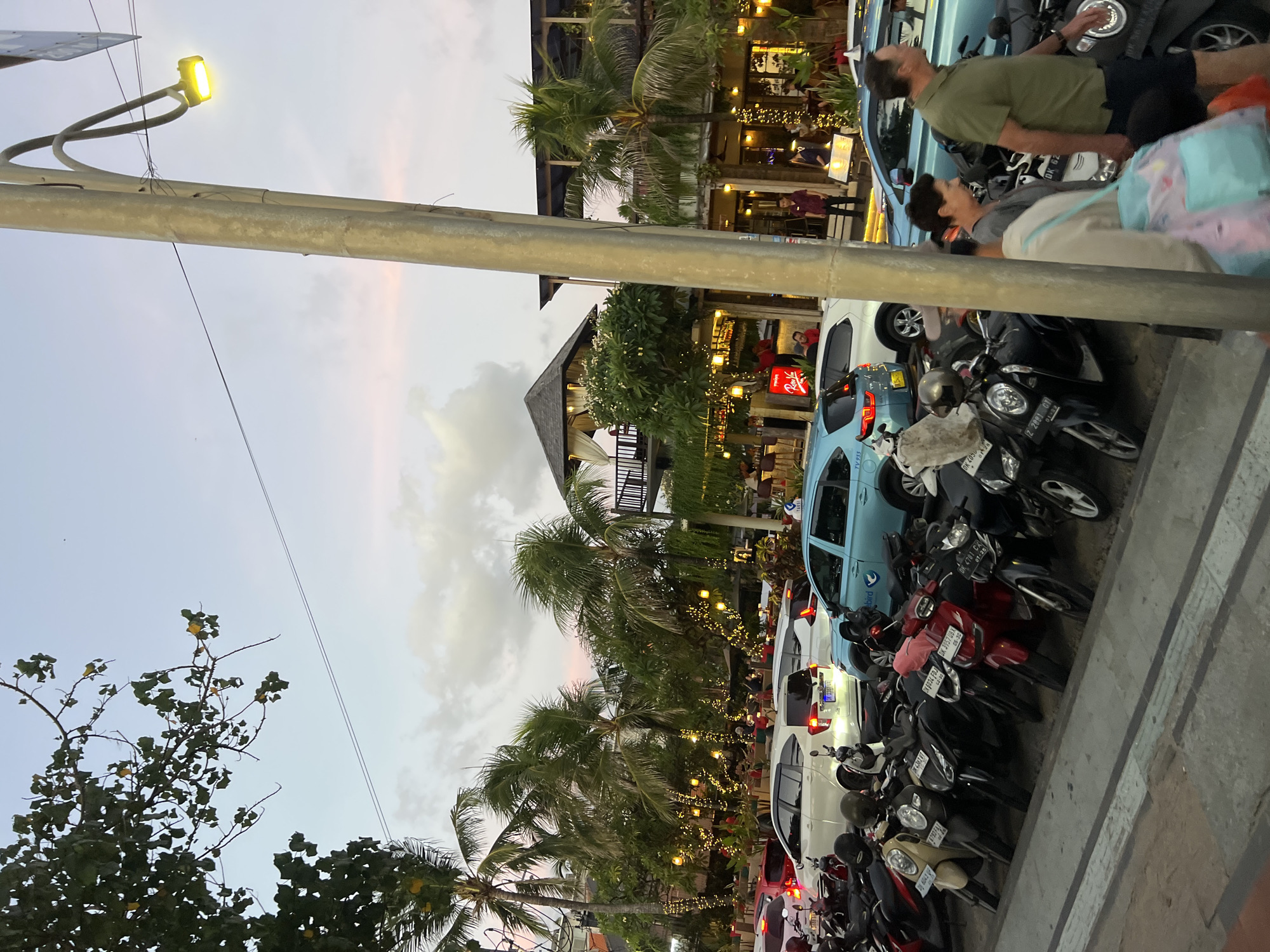 In Kuta, Indonesia, on a street lined with palm trees, a parking area is filled with numerous motorcycles and several cars. The scene appears to be from dusk, with a soft, pastel-colored sky. Buildings housing shops and restaurants with outdoor seating are visible behind the parked vehicles. String lights are illuminated, creating a festive atmosphere. A street lamp stands prominently in the foreground, casting a warm glow. A few people are walking by, some with bags, suggesting activity and commerce. The overall impression is a lively urban environment at the end of the day.
