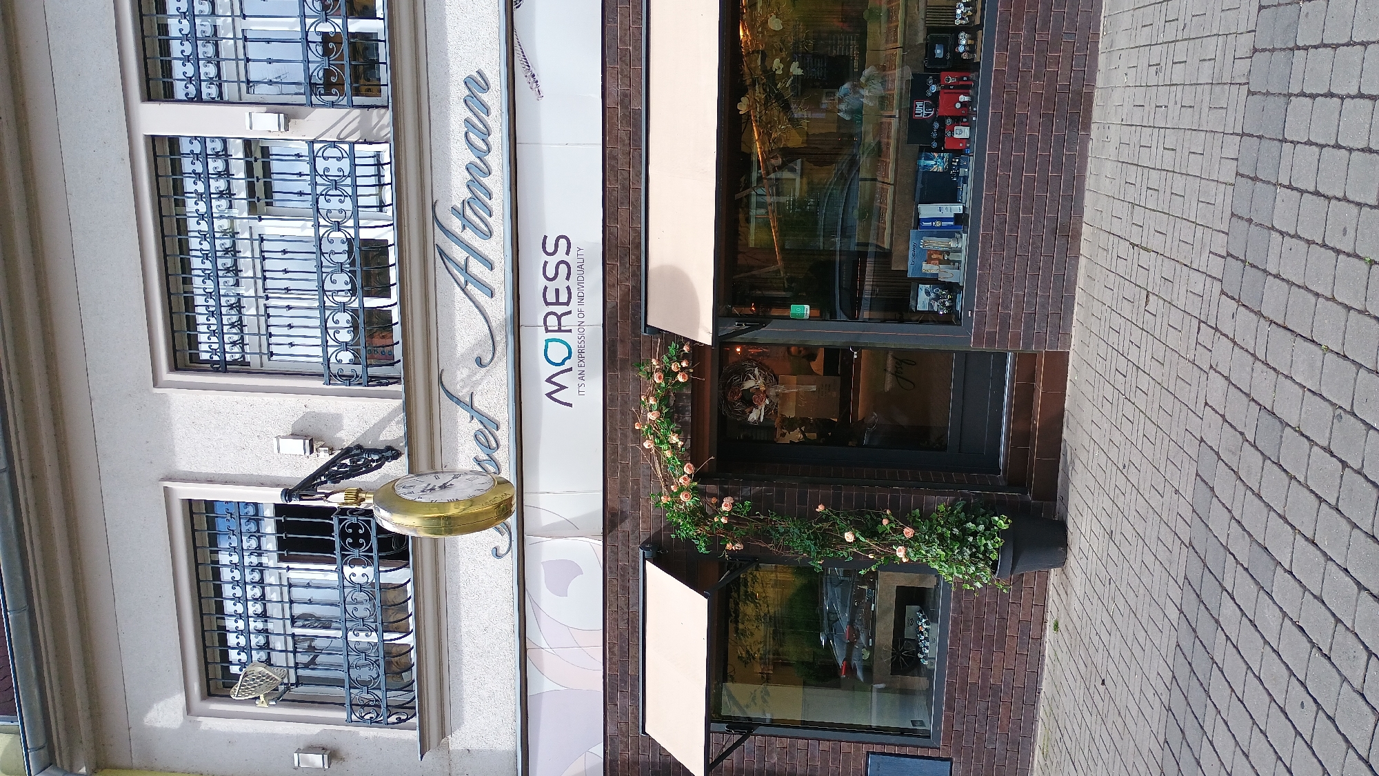 The image shows the exterior of a shop in Sokolov, Czechia. The shop's name, "Josef Altman," is visible in ornate script above the storefront, alongside the word "MORESS" and the tagline "IT'S AN EXPRESSION OF INDIVIDUALITY." A large, ornate pocket watch hangs as a sign above the entrance. The storefront features large display windows showcasing various watches and other items. A potted plant with pink flowers and greenery is positioned next to the entrance. The building has windows with decorative iron grates on the upper level. The ground in front of the shop is paved with cobblestones. The lighting suggests it is daytime.