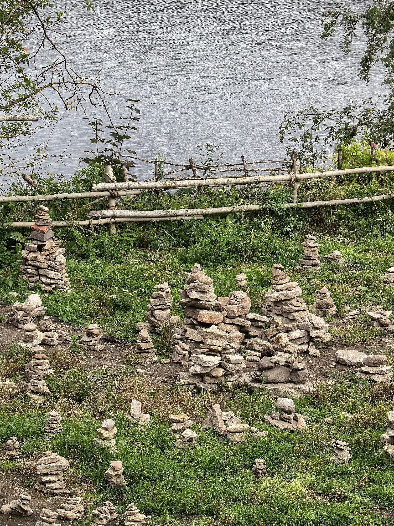 The image depicts a landscape in Staraya Ladoga, Russia, characterized by a body of water in the background, separated from the foreground by a rustic wooden fence. The foreground is a grassy slope dotted with numerous cairns, or stacks of stones. These cairns vary in size and composition, with some appearing to be carefully constructed and others more haphazard. The stones are predominantly gray and brown, reflecting the natural geological material of the area. The environment is verdant, with lush green grass and some interspersed dry vegetation. The overall scene suggests a natural, possibly historical or spiritual site, where visitors have engaged in the practice of stacking stones.