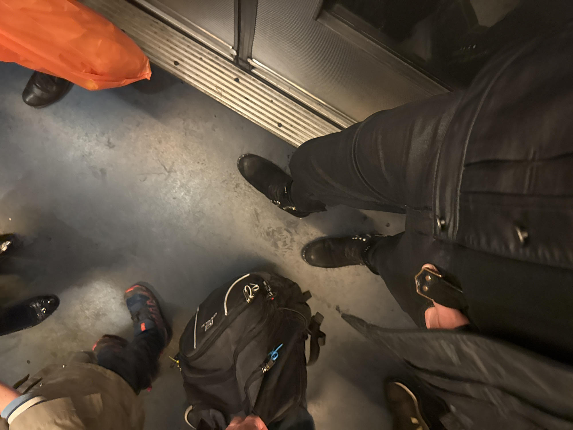 This image appears to be taken inside a train in Paris, France. The scene shows the lower portion of several people standing inside the train, with a focus on their feet and belongings. An orange plastic bag, black dress shoes, a backpack, and various boots are visible on the train floor. The train's interior includes a ridged floor and metal door trim.