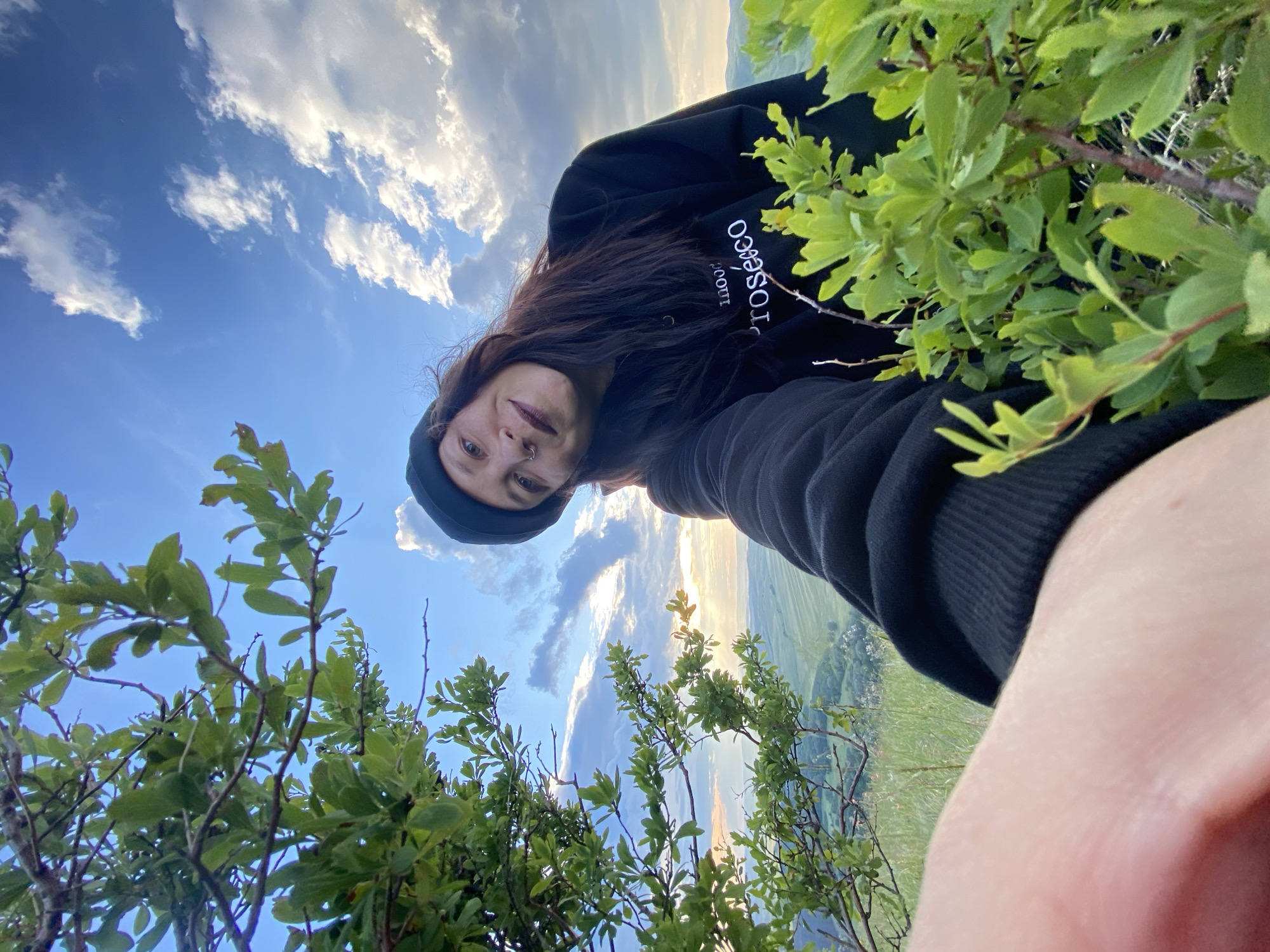 A medium shot captures a woman taking a selfie outdoors in a grassy area with a scenic landscape visible in the background. The woman is wearing a black beanie and a black sweatshirt with white text that reads "Prosecco." Her arm, also clad in black, extends towards the camera. She has dark hair, a nose ring, and is smiling. Lush green foliage frames the foreground on the left and right sides of the image. The sky above is blue with scattered white clouds. The background shows rolling hills, fields, and trees, suggesting a rural or natural setting. The location is identified as the city of Абзаново, Russia.