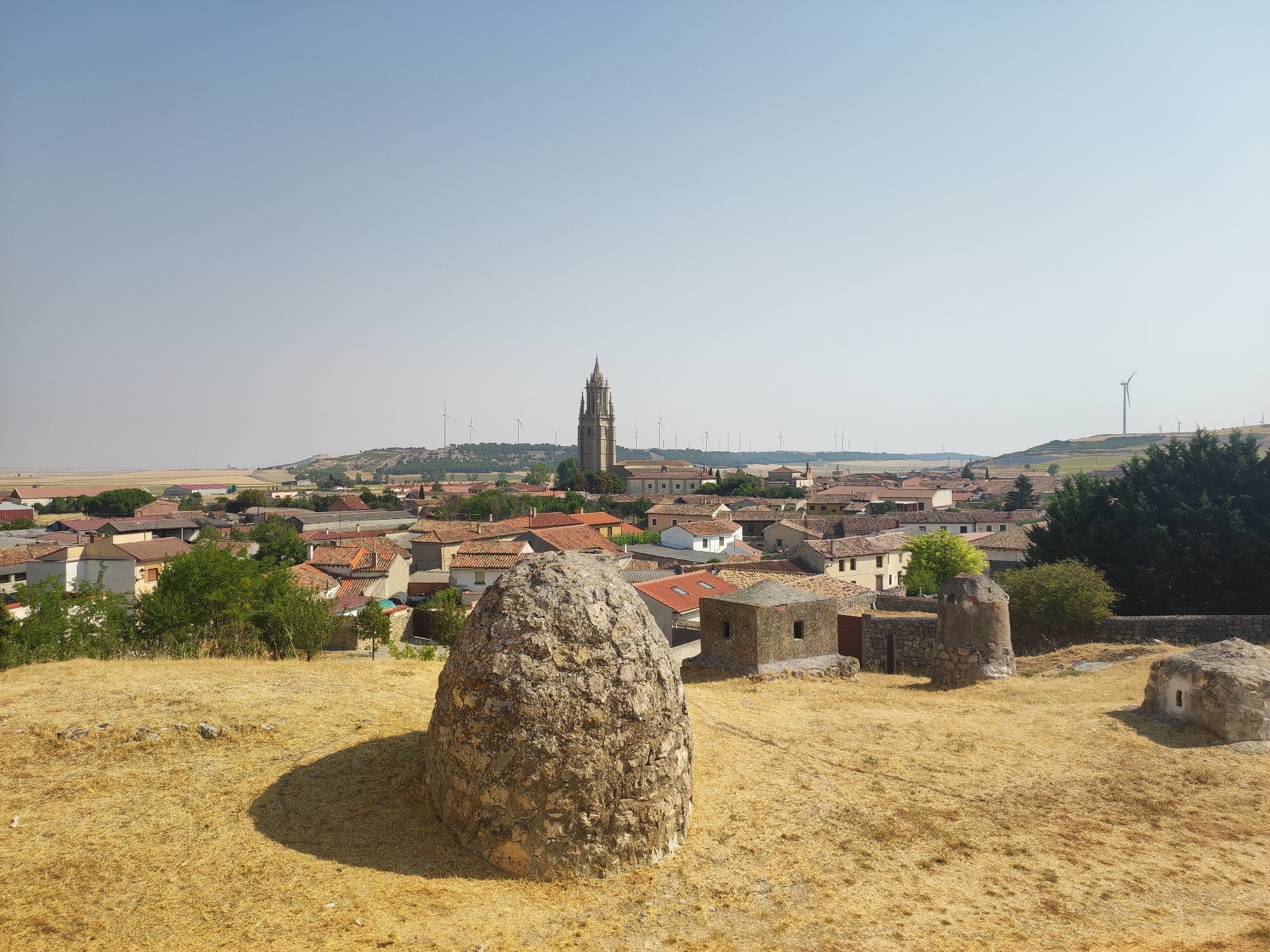 The image shows a panoramic view of the city of Ampudia, Spain, on a clear day. In the foreground, there is a large, rough, egg-shaped stone structure casting a shadow on the dry, golden grass. To the right, there are two smaller stone structures, one resembling a miniature watchtower and another a small, cubical structure with an opening. Beyond these structures, the town of Ampudia stretches out with its characteristic terracotta-tiled roofs. A prominent church tower with a Gothic spire rises above the buildings in the middle ground. The background features rolling hills dotted with numerous wind turbines. The overall landscape is arid, with dry vegetation and a pale blue sky. There are no people visible in the image.