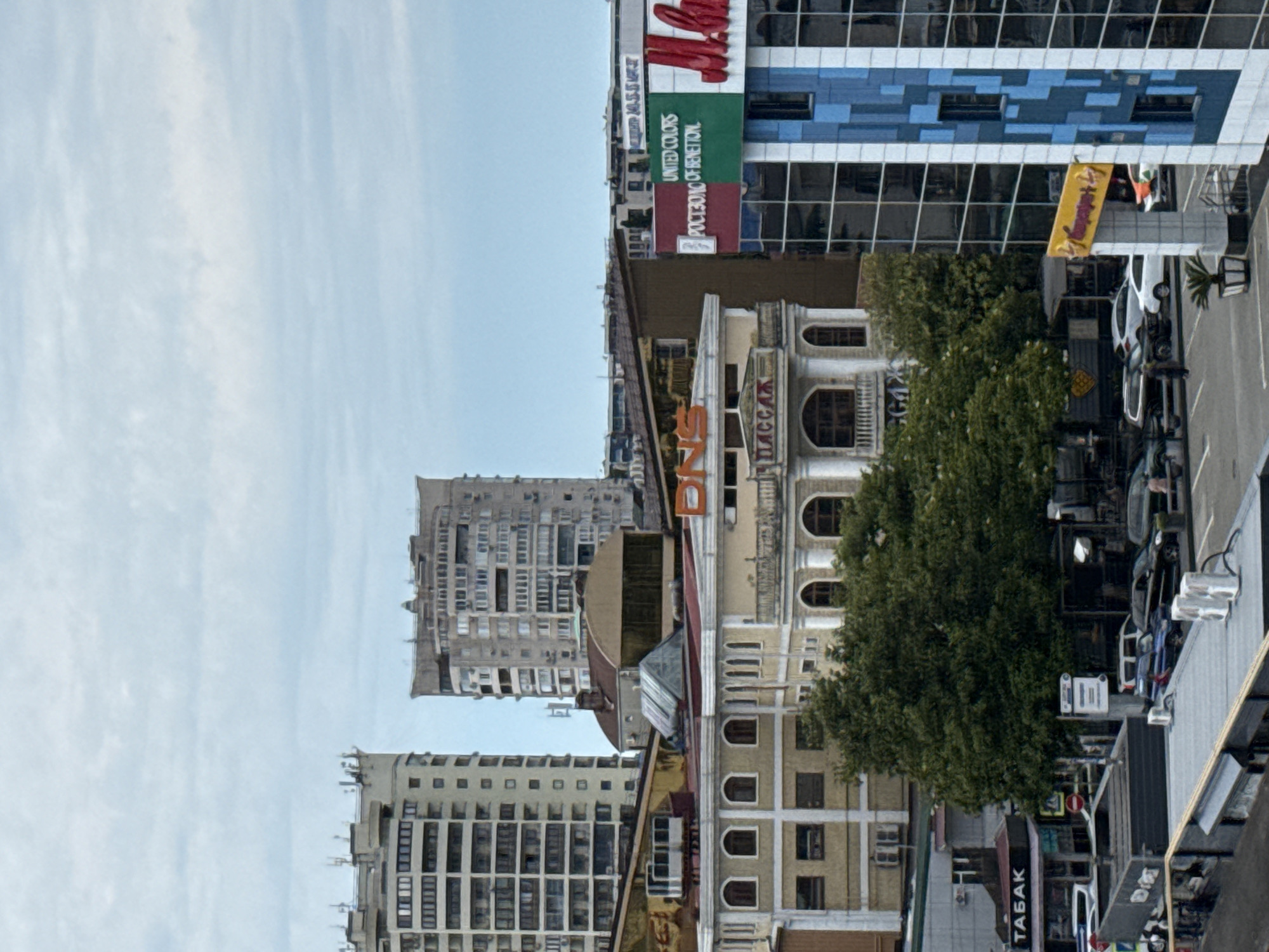 The image depicts an urban landscape in Sochi, Russia, under an overcast sky. In the background, several tall apartment buildings are visible, characterized by their multi-story facades and numerous windows. One building, in particular, has a light beige color and a distinct tiered structure.
In the mid-ground, a cream-colored building with arched windows and decorative balconies is prominent. Above this building, a sign with orange lettering reads "DNS." Another sign below it has the word "ПАССАЖ" in white letters on a dark background. To the right of this structure, a modern building with a glass facade displays a green and red sign that reads "UNITED COLORS OF BENETTON" and partially visible text in red.
The foreground shows a street scene with parked cars and pedestrian activity. A sign with the Cyrillic word "ТАБАК" (Tobacco) is affixed to a storefront. Several trees, with dense green foliage, are interspersed throughout the scene, providing a contrast to the architectural elements. The overall impression is a bustling city environment with a mix of residential, commercial, and retail spaces.