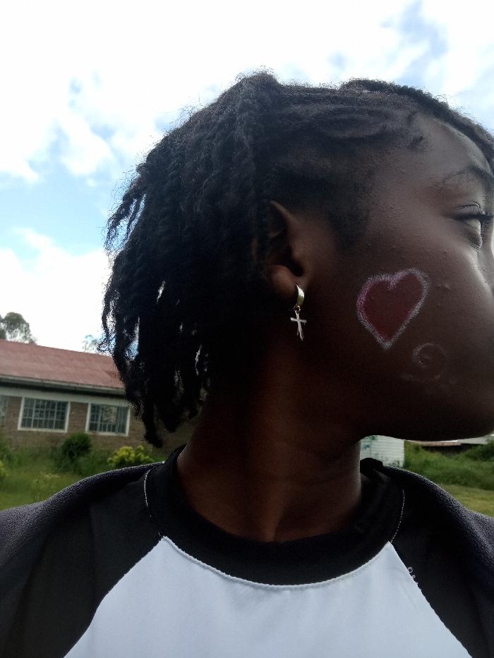 A person with dark skin and braided hair is seen in a close-up shot. They are wearing a silver cross earring. A heart-shaped design in red is painted on their cheek. They wear a black and white raglan shirt. The background includes a building with multiple windows and some greenery, suggesting an outdoor setting beneath a sky with scattered clouds.