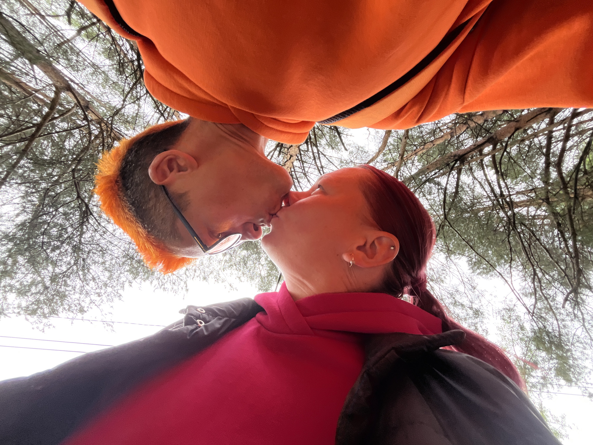 A low-angle, close-up shot depicts two individuals kissing beneath a canopy of trees. The individual on the left has short, vibrant orange hair and wears glasses and an orange hoodie. The individual on the right has long, reddish-brown hair pulled back in a ponytail and is wearing a pink hooded sweatshirt and a black jacket. Both individuals are facing each other, with their lips touching in a kiss. The background features a dense network of tree branches against a gray sky. The location is Chaikovsky, Russia, based on the EXIF data.