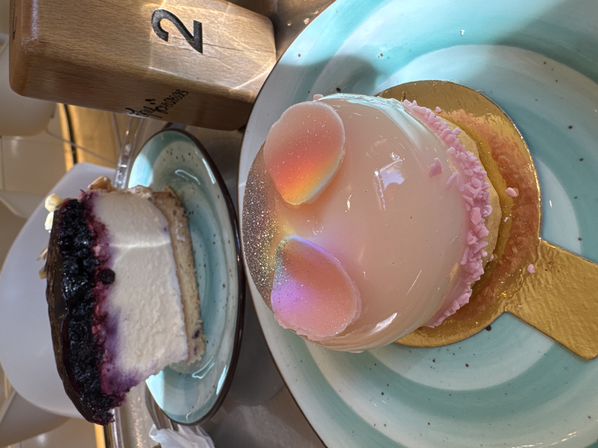 A close-up, eye-level shot shows two desserts on teal-colored plates. The dessert in the foreground is a round, light pink mousse cake with a glossy finish, topped with edible glitter and two pink, heart-shaped decorations. The cake sits on a gold base and is decorated with pink sprinkles around its bottom edge. The background features a slice of blueberry cheesecake on a similar teal plate. To the right, a wooden table number "2" is visible. The setting appears to be a cafe or restaurant. The overall lighting is bright and natural. This scene could be located in Moscow, Russia.