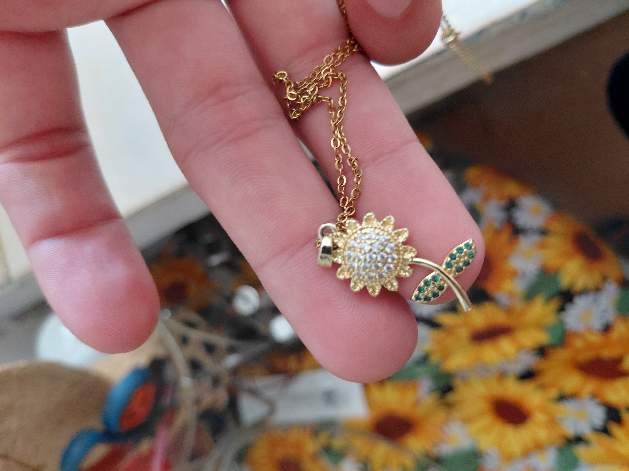 A light-skinned human hand is centrally positioned, holding a gold-colored necklace featuring a sunflower-shaped pendant. The pendant's central disk is densely set with small, clear, circular-cut gemstones, surrounded by gold-colored petals. Below the sunflower head, a slender gold-colored stem extends downwards, bearing two leaf-like elements, each meticulously set with multiple small, green, circular-cut gemstones. The necklace chain is delicate and gold-colored, draping loosely from the pendant. The background is significantly out of focus; the lower half displays a blurred pattern of yellow and brown, consistent with sunflowers, alongside some lighter, possibly white, floral patterns on a textile surface. The upper left background contains indistinct, blurred shapes in various colors, including brown, blue, and red, suggestive of other objects in a cluttered environment. The location is unknown.