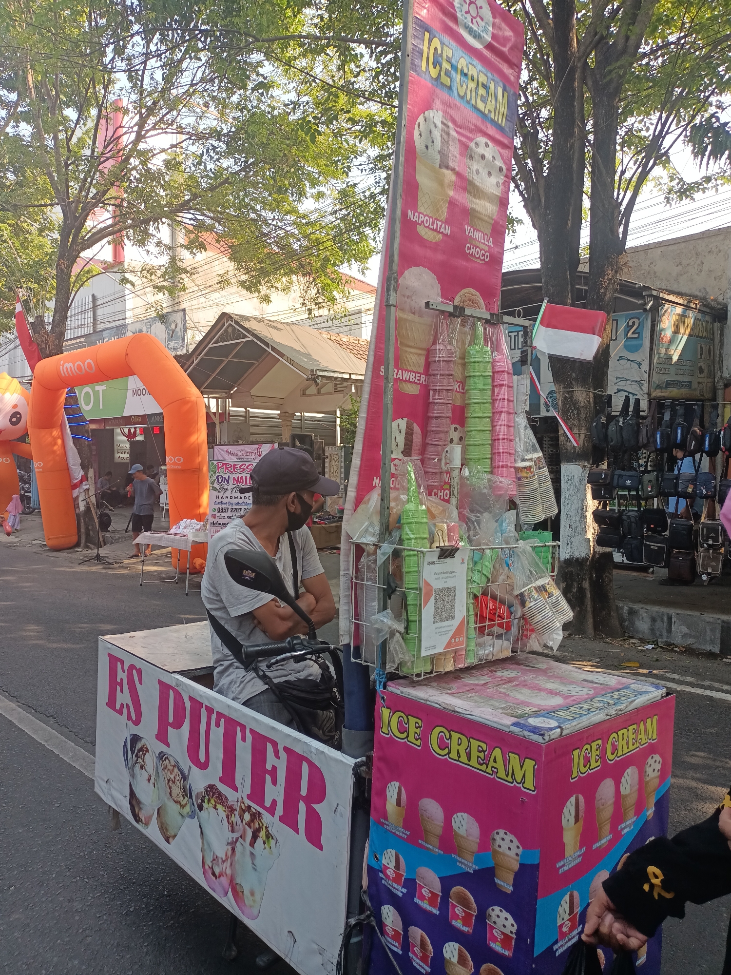 A street vendor is operating a mobile ice cream cart in Pare, Indonesia, on a sunny day. The vendor, wearing a grey t-shirt, dark cap, and black face mask, sits on a motorcycle that powers the cart. The cart is adorned with colorful banners advertising "ICE CREAM" and "ES PUTER," featuring images of ice cream cones and cups. Various ice cream cups, cones, and toppings are displayed on the cart. In the background, an orange inflatable arch with the "imo" logo stands, and several shops line the street. People are visible in the background, some browsing merchandise and others attending to activities near the arch. The street is paved, and trees with lush green foliage provide shade. The overall scene depicts a bustling street market environment in a commercial area.