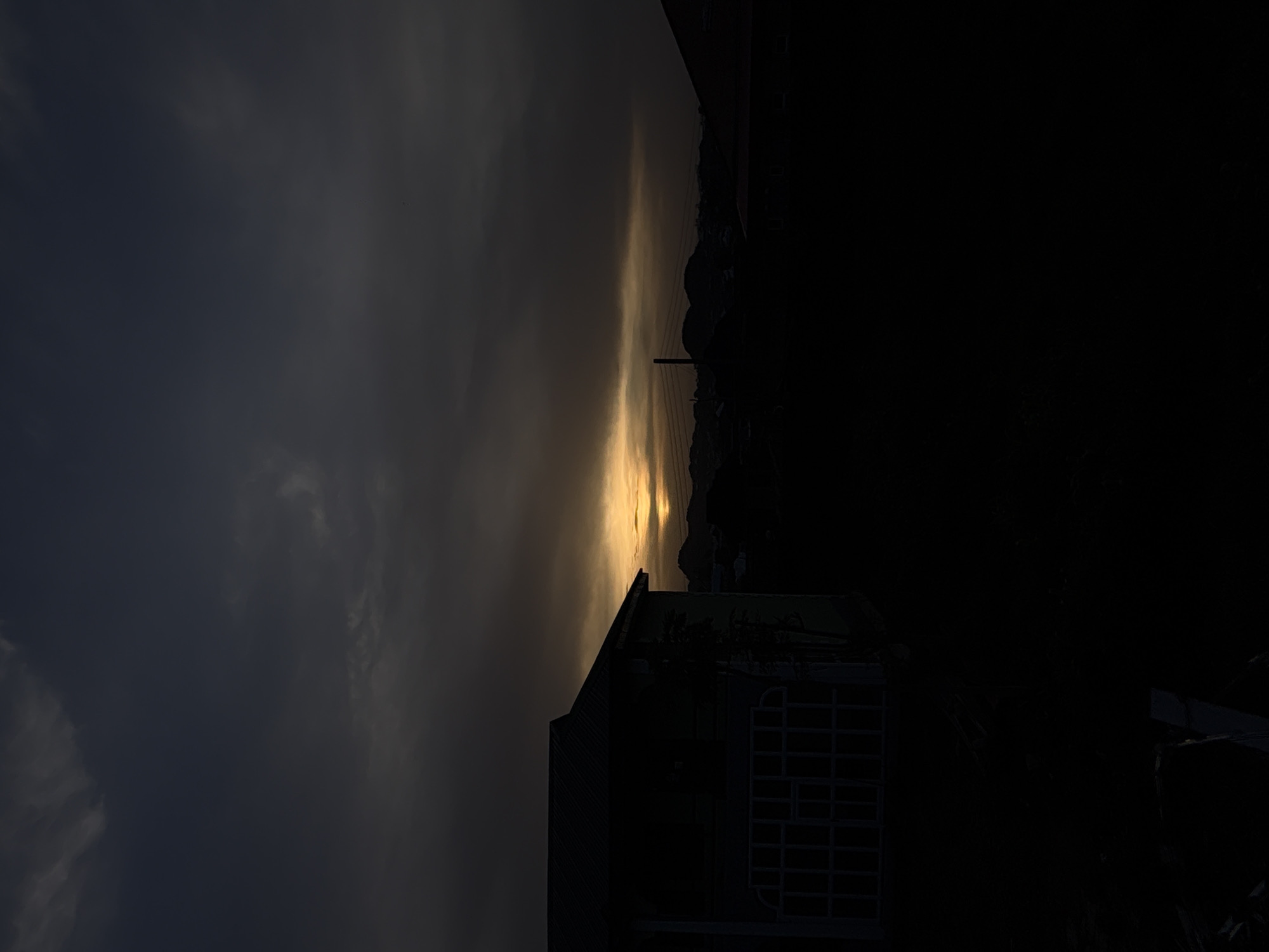 The image, when correctly oriented, depicts a dramatic sunset or sunrise over a landscape, with most elements rendered in silhouette. The sky dominates the upper portion, featuring heavy, dark grey and blue-grey clouds that transition towards the horizon.
At the mid-ground, a brilliant, horizontal band of golden-yellow light pierces through the clouds, marking the position of the sun, either rising or setting. Against this bright horizon, a range of silhouetted hills or mountains is visible. A tall, slender utility pole with multiple horizontal lines (likely power lines) stands prominently in the mid-ground, slightly to the right of the center.
The foreground is almost entirely plunged into darkness, presenting an array of silhouetted structures. On the left, the dark, angular roofline and upper portion of a building are visible. To the right of this building, a complex, dark, grid-like structure, possibly a large window frame, a section of a fence, or part of a building's facade, stands out against the dim light. The very bottom right portion of the image is completely black, indicating deep shadow or an obscured area.
The overall scene conveys a somber yet striking mood due to the strong contrast between the radiant horizon and the dark, featureless foreground. No discernible people or visible text are present in the image. The heavy clouds suggest an overcast or partially cloudy day, with the sun breaking through close to the horizon. The context provided indicates the location is Bukuru, Nigeria.