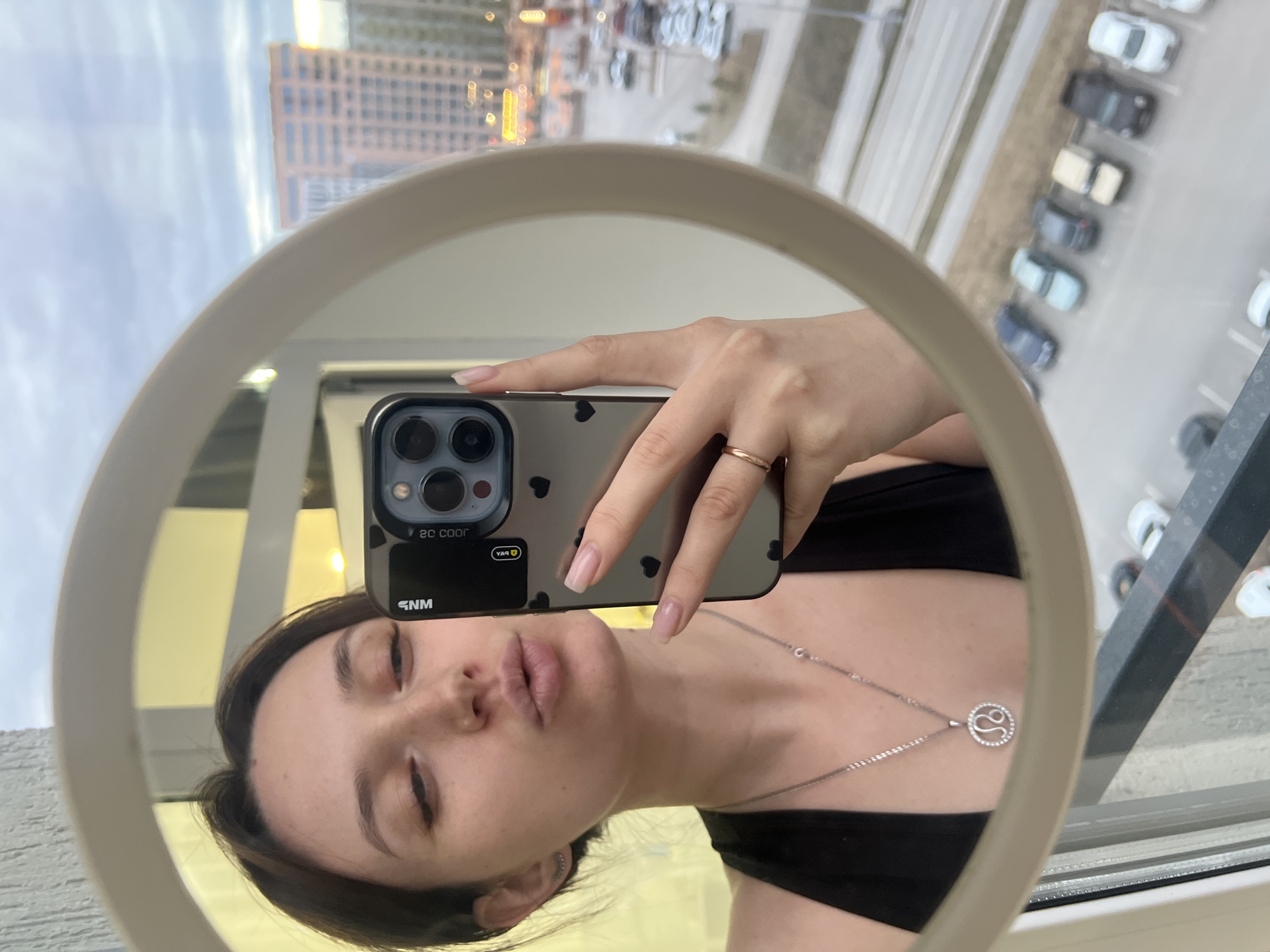 The image displays a mirror selfie of a young woman, captured within a round, light-colored mirror, and is rotated 90 degrees counter-clockwise.
The primary subject, a woman, is seen from the chest up, reflected in the mirror. She is wearing a black top and a silver-colored necklace featuring a circular pendant, which appears to be the Leo zodiac symbol, adorned with small gems. Her left hand holds a smartphone, likely an iPhone, which is also reflected. The phone is encased in a clear cover decorated with small black heart decals. A simple gold-colored ring is visible on her left ring finger. Her face shows closed or partially closed eyes and her lips are in a slight pout.
Text visible on the phone's screen reflection includes "20 COOR" and "5NM", both appearing upside down due to the mirror and phone orientation.
The background, visible around and above the mirror's reflection, is an urban landscape seen through a window. The sky is overcast, and several multi-story buildings are visible, one reflecting warm yellow light. Below the buildings, a paved parking lot is discernible, featuring multiple cars parked in rows, mostly light-colored. The scene suggests an elevated perspective, possibly from a high-rise building in Tyumen, Russia, during daytime.