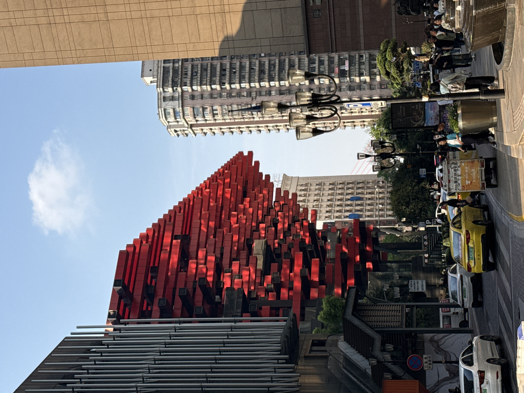 The image shows a street scene in the Yuzhong District of Chongqing, China. The most striking feature is a large, modern building with a facade constructed from numerous red and black rectangular blocks. A metal structure composed of parallel horizontal bars is located in the foreground, partially obscuring the base of the red building.
In the background, there are several other taller buildings, with a light-colored, grid-patterned building visible behind the red structure.
The right side of the image shows a street with vehicles parked and moving along the road. Cars, including a yellow one, are parked on the side of the street. Pedestrians are also present. Street lights are also visible.