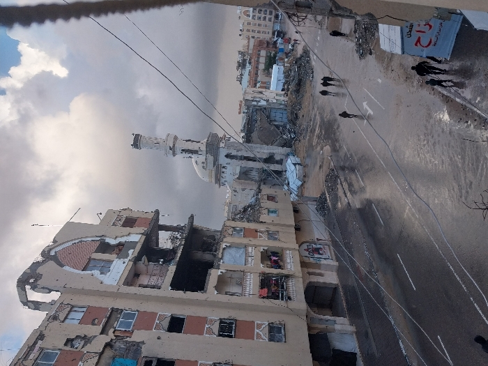 The image captures a severely damaged urban landscape in Khan Yunis, Palestinian Territory, under an overcast sky. A multi-story residential building on the left exhibits extensive structural damage, with entire sections of walls and floors missing, exposing internal rooms and balconies. Debris is visible on and around its lower levels. In the middle distance, a mosque stands with a heavily damaged minaret, its top section destroyed, and visible damage to its dome and main structure. The paved street in the foreground and mid-ground is wet and reflective, indicating recent rain or standing water, with debris scattered along its edges. Approximately seven individuals are walking on the street and sidewalks; a man and a child are visible on the sidewalk in the lower right, while a group of three to four people walks in the center of the wet street. Further back, more collapsed structures and piles of rubble are present, along with what appear to be temporary shelters or tents. Overhead wires traverse the scene diagonally.