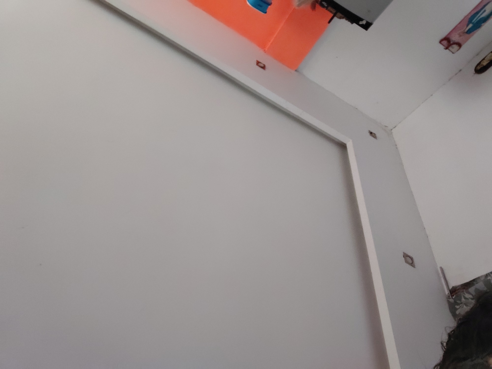 The image depicts an indoor scene, likely a room, characterized by white painted walls and ceiling. A white architectural or furniture element, possibly a frame or beam, traverses the upper and right edges of the primary wall. Along this white structure, several small, metallic, square plates with circular cutouts are visible, potentially serving as fasteners or decorative elements.
In the upper central portion of the image, a bright orange object, which could be a shelf or a box, is partially visible above the white frame. A small, indistinct blue item rests on this orange surface. The top right corner of the room features some casual decorations, including a blurred object that appears to be a sticker or small drawing with blue and pink hues.
The top of a person's head, with dark hair, is cropped into the bottom right corner of the frame. No specific activity is occurring, and the scene appears static. The lighting is diffused, highlighting the white surfaces. No legible text is discernible within the image. The setting suggests a casual, possibly domestic, interior environment.
