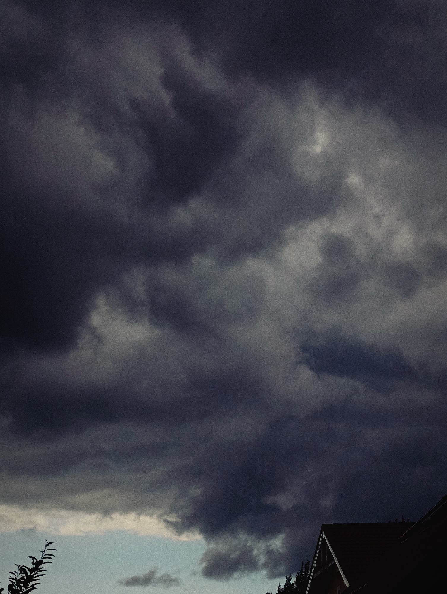 The image shows a dark, cloudy sky over a residential area. The clouds are heavy and ominous, suggesting an approaching storm. In the lower right corner, the roof of a house is visible, with its dark shingles contrasting against the lighter sky. A silhouette of a tree branch is present in the lower left corner. The overall mood of the image is dark and dramatic, with a hint of unease. The location is identified as Unknown, Unknown.