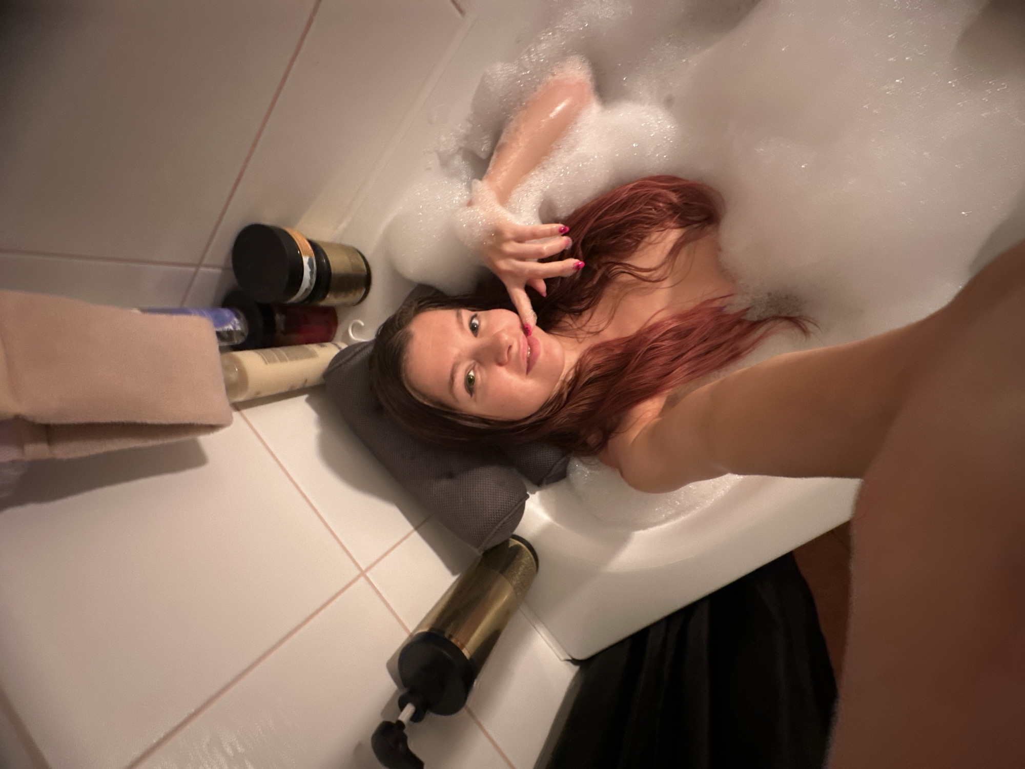 A woman with reddish-brown hair is in a bathtub filled with bubbles. She is lying on a gray pillow. Beside the bathtub, on the tiled floor, are several bottles and jars, as well as a stack of towels. A black cloth hangs from the side of the tub. The location is unknown.