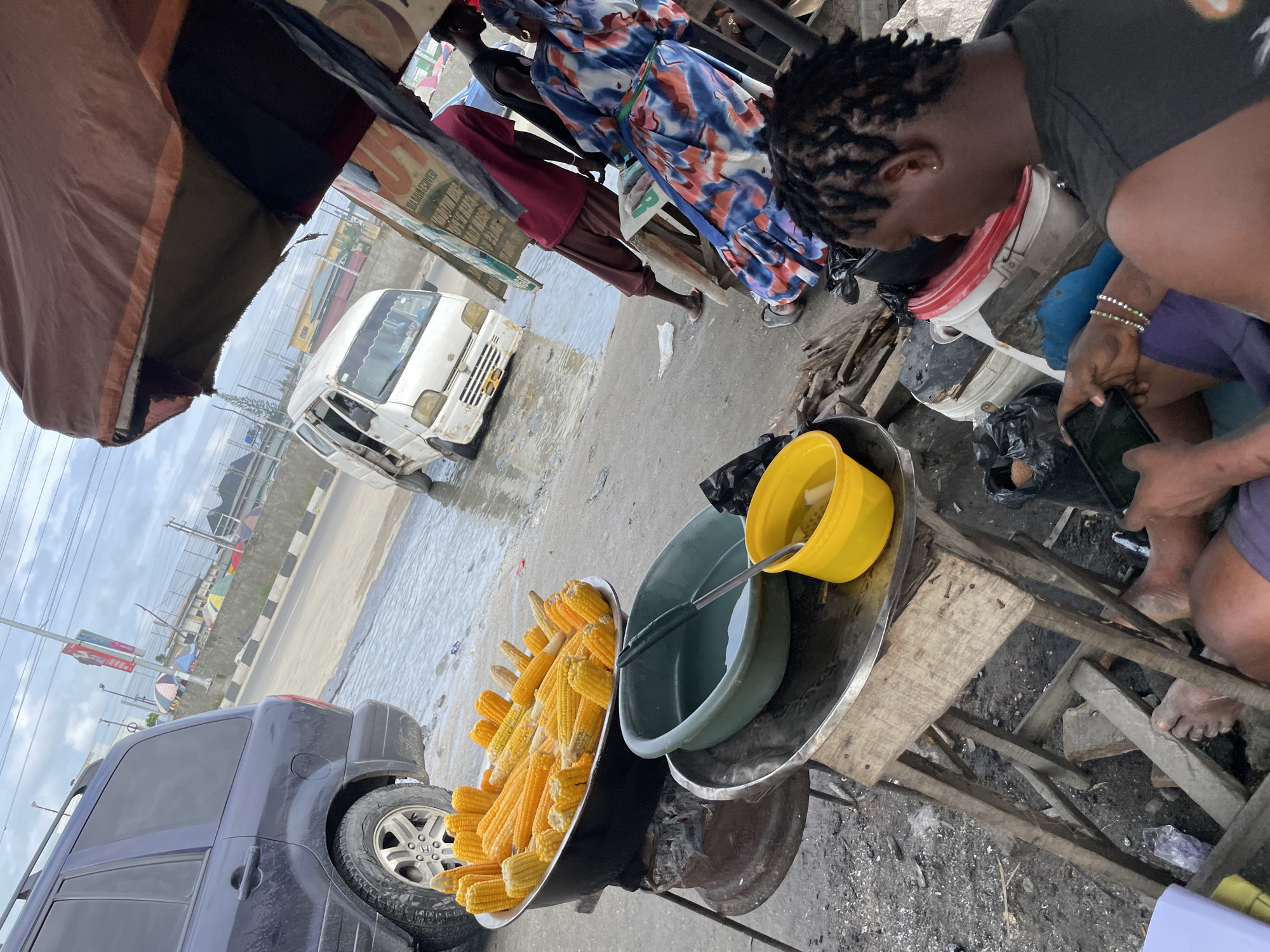 The image depicts a street scene in Lekki, Nigeria. The primary subjects are two individuals, one of whom is a young woman with braided hair, dressed in a dark t-shirt and purple shorts, engrossed in using a mobile phone. She is seated on what appears to be a makeshift wooden stool. The other person, partially visible, is wearing a colorful blue and red patterned dress and maroon pants.
The scene is a bustling marketplace or roadside stall. Prominently featured are large metal bowls filled with corn on the cob, suggesting that corn is being sold or prepared. A large green basin contains water and a ladle, likely for washing or cooking. A yellow bucket is also present. In the background, a white van is parked on a wet, possibly flooded, road. A dark-colored SUV is also visible on the left side of the frame. The overall setting suggests an outdoor environment, with a tent or awning providing shade.
The activity appears to be the daily commerce of street vending. The presence of water and the general atmosphere hint at a tropical climate, and the wet ground could indicate recent rainfall or a generally humid environment. There are no clear indications of the specific time of day, but the lighting suggests it is daytime. The image captures a candid moment of everyday life in this Nigerian location. While some text is visible on a sign in the background, it is not legible enough to be described.